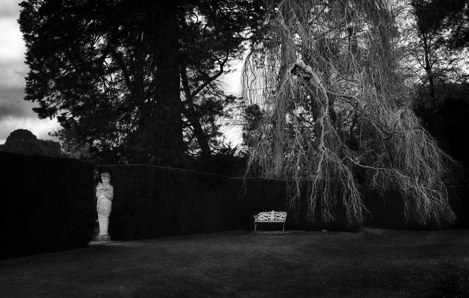 A black and white photo of a garden with a statue of Pan, a bench in the middle, and leafless trees in the background.