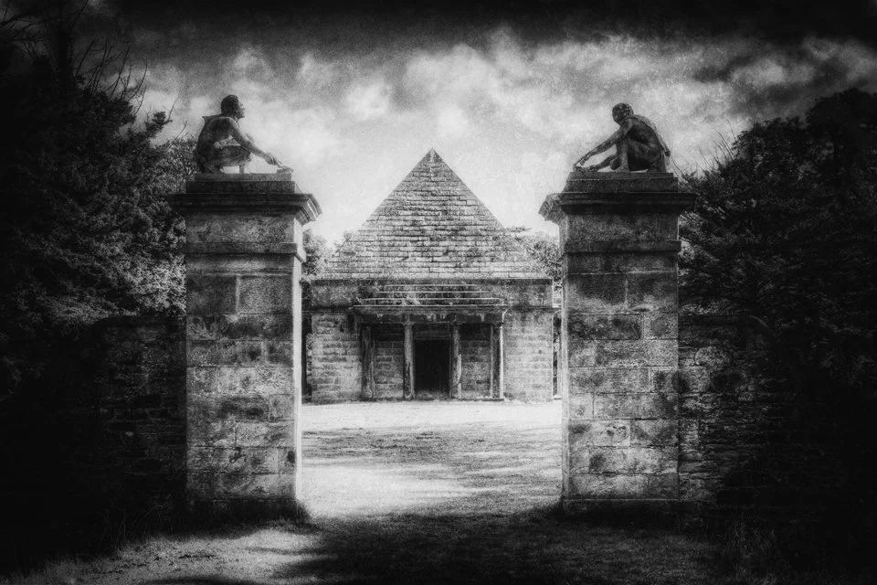 Black and white photo of the Wemyss Pyramid Mausoleum  at Gosford Park,. Two stone statues of seated figures, one on each of the front pillars, look over the entrance. Surrounding trees frame the scene under a cloudy sky.