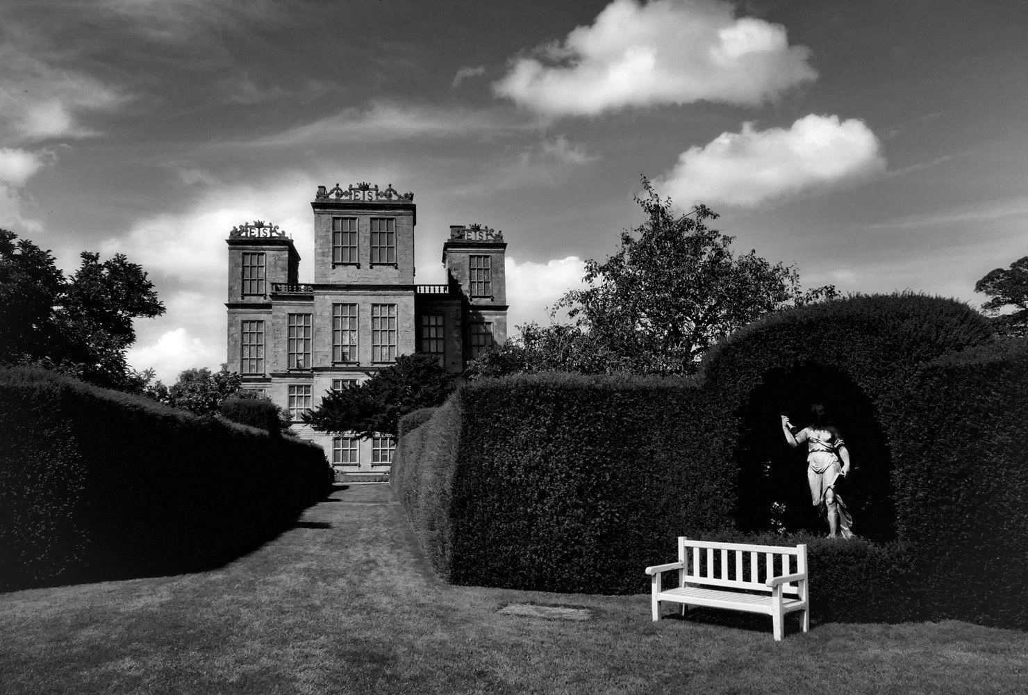 A black-and-white photo of  the garden at Hardwick Hall, with trimmed hedges, a statue of a woman in deep shadow, and a white bench in front of the statue. 