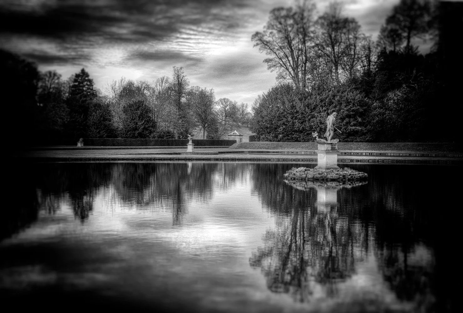 Black and white photo of the water gardens at Studley Royal with a statue, reflecting in a still pond surrounded by trees and cloudy sky.