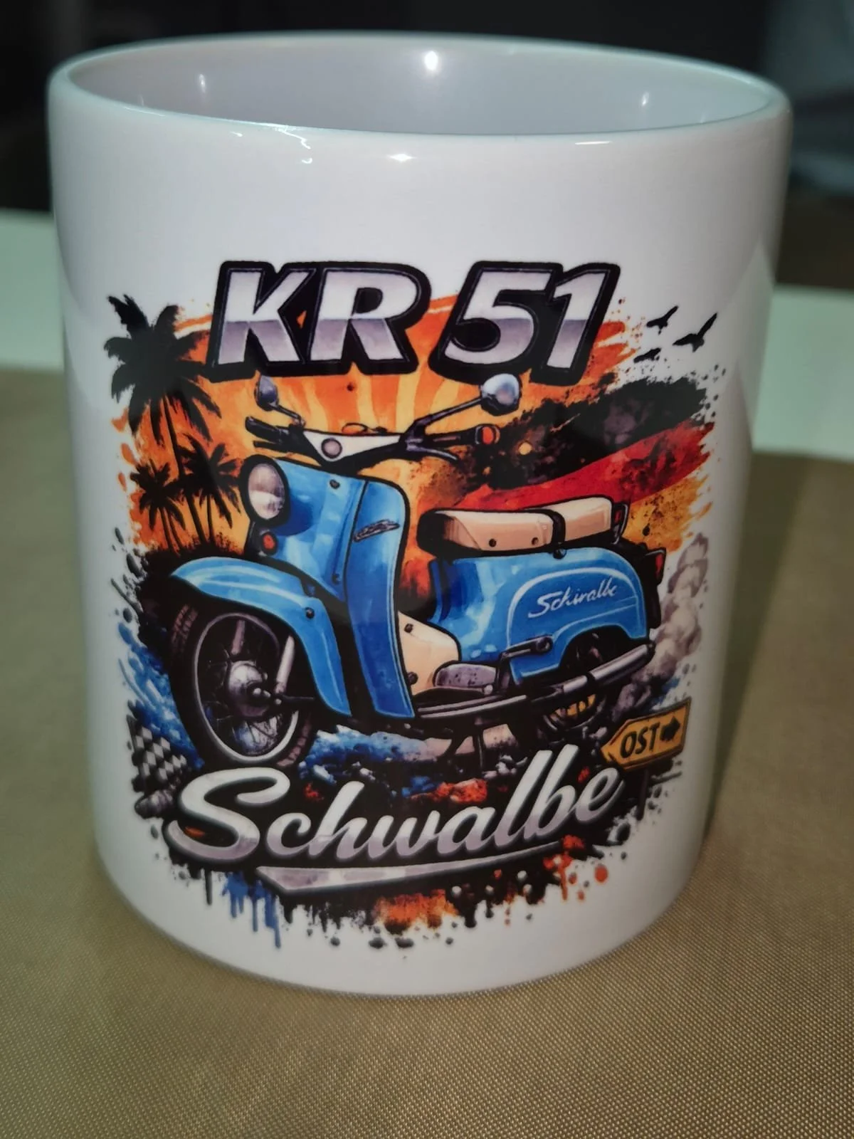 Tasse mit einem bunten, graffitiartigen Design einer blauen Simson Schwalbe Kr51, Palmbaum, Sonnenuntergang im Hintergrund, sowie den Texten 'KR 51', 'Schwalbe' und einen Wegweiser mit der Aufschrift 'Ost'.