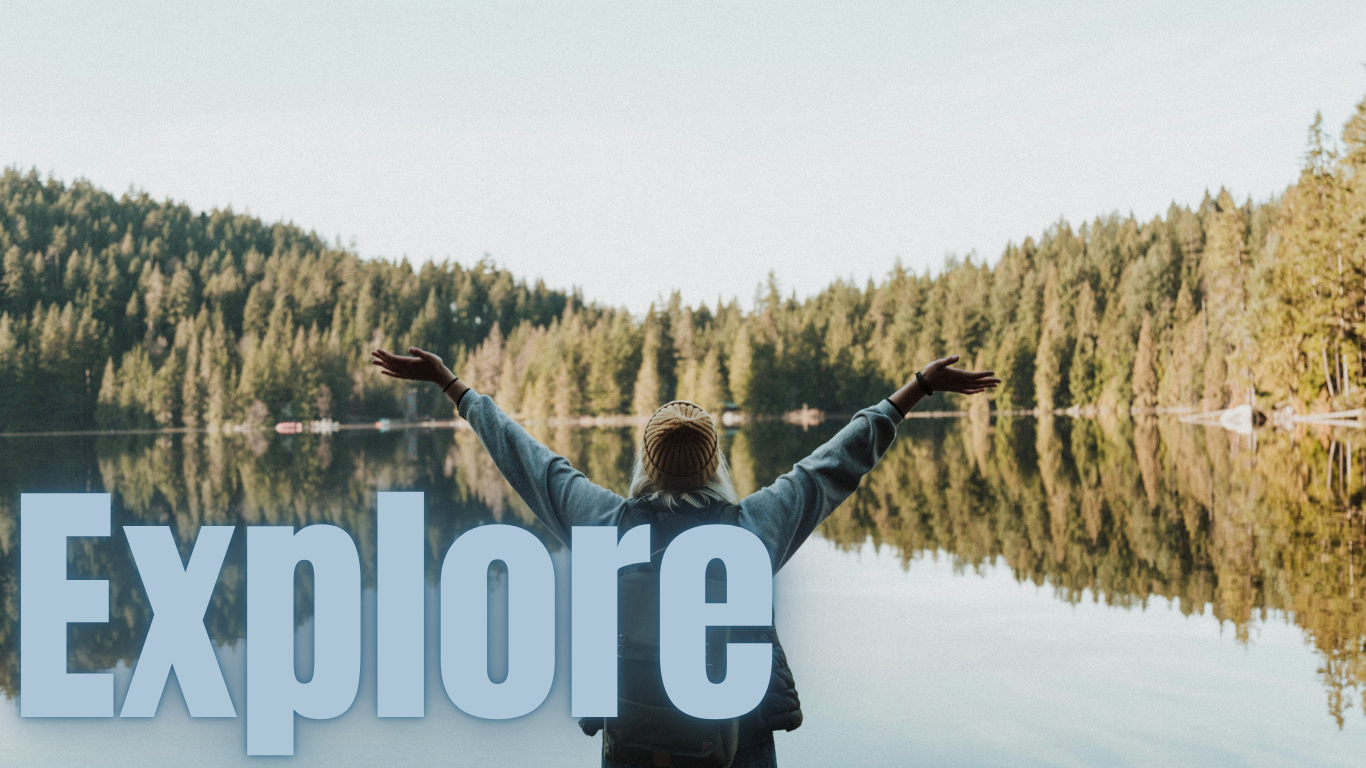 Person in gray hoodie and brown beanie with arms raised, standing in front of a calm lake with forested shoreline, reflected in the water, with the word 'Explore' in large blue letters.