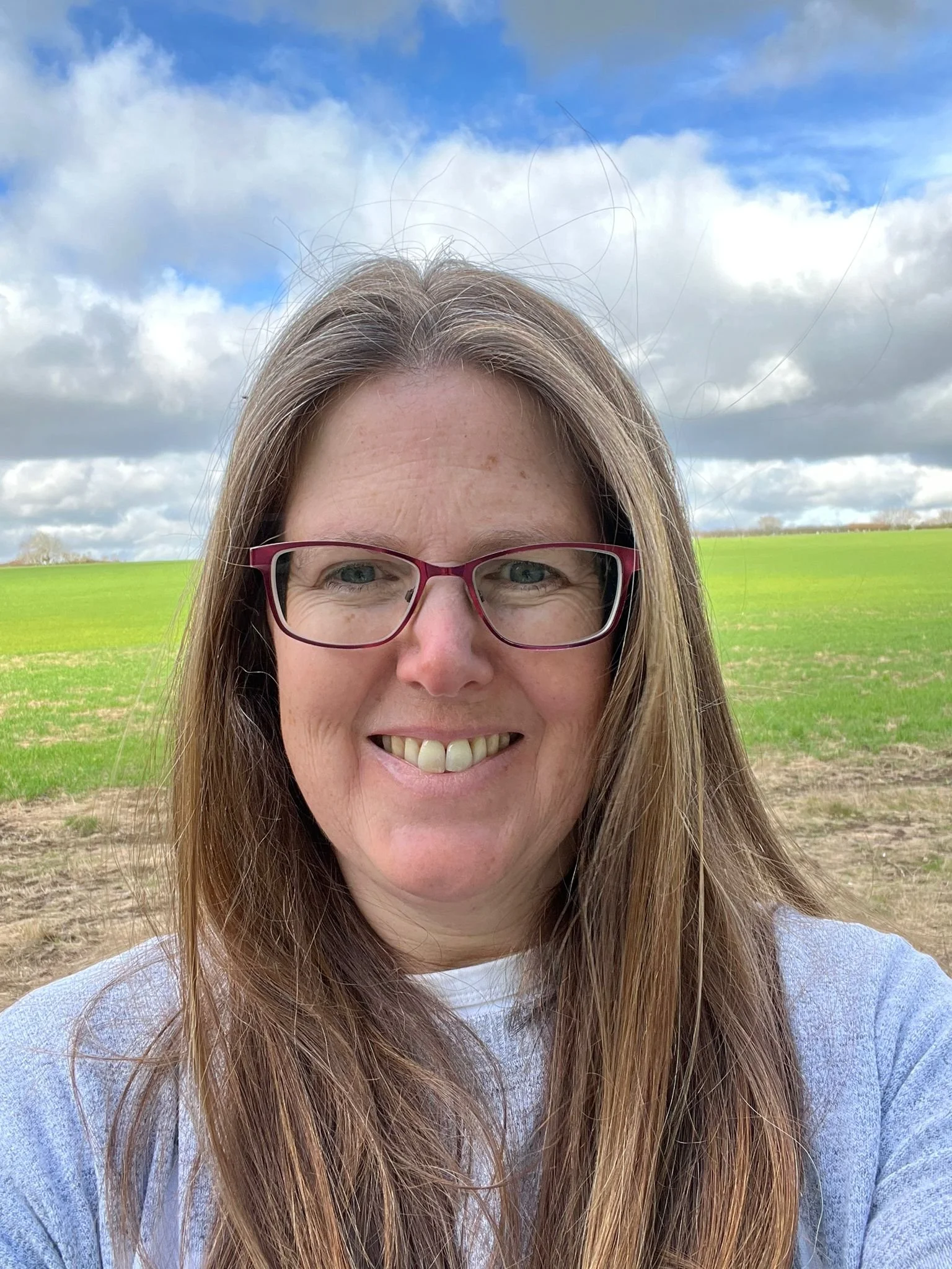 Close-up of a woman with long brown hair and glasses smiling outdoors, with a green field and a cloudy blue sky in the background.