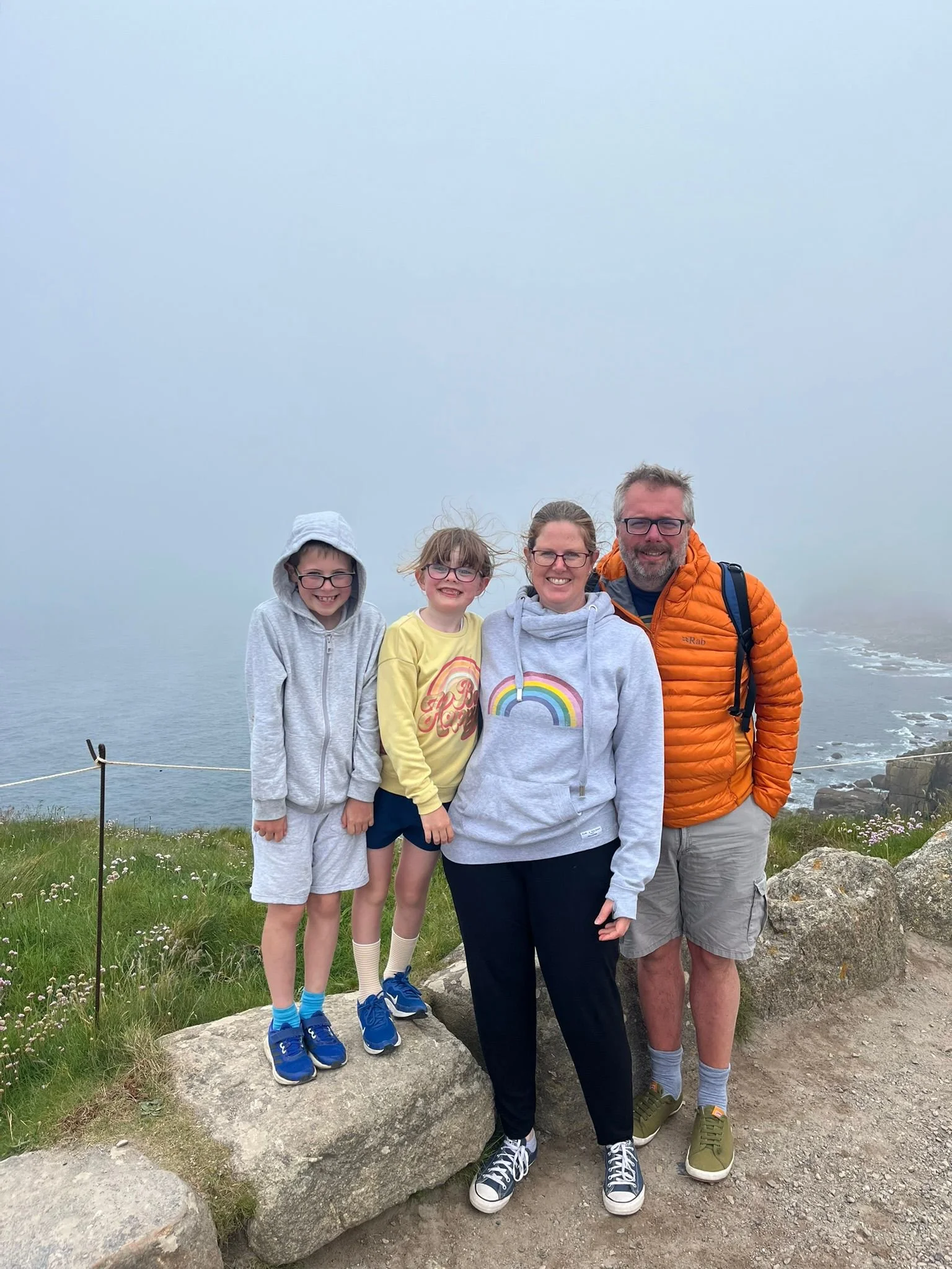 A group of four people, two children and two adults, standing outdoors on rocky terrain near the water with a foggy sky, smiling at the camera.