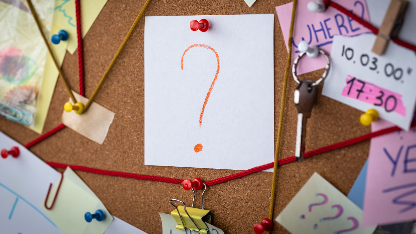 A cork bulletin board decorated with colorful notes, pins, and a key. A white paper with a large orange question mark is pinned in the center.