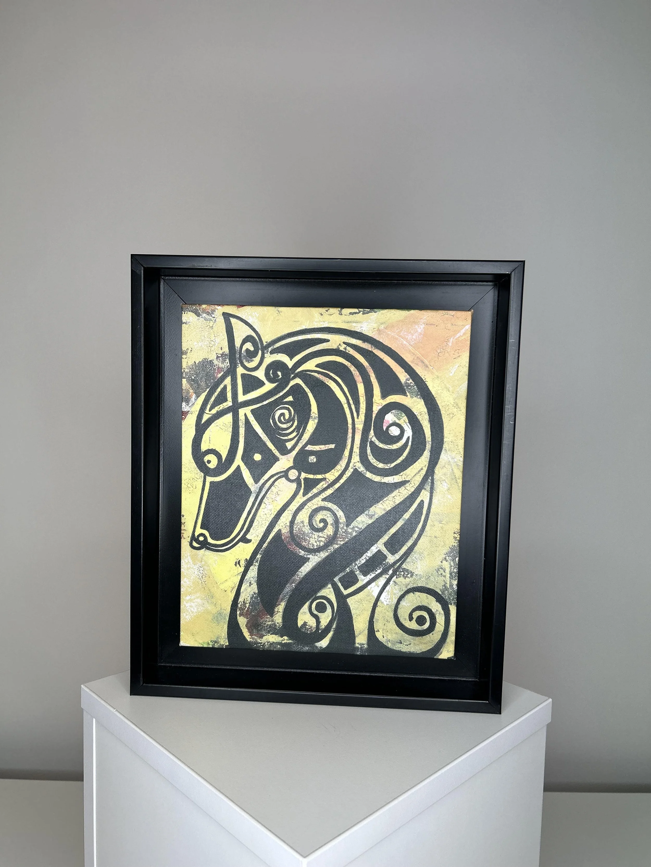 Framed artwork featuring a black abstract curvilinear design on a yellowish background, displayed on a white pedestal against a plain gray wall.