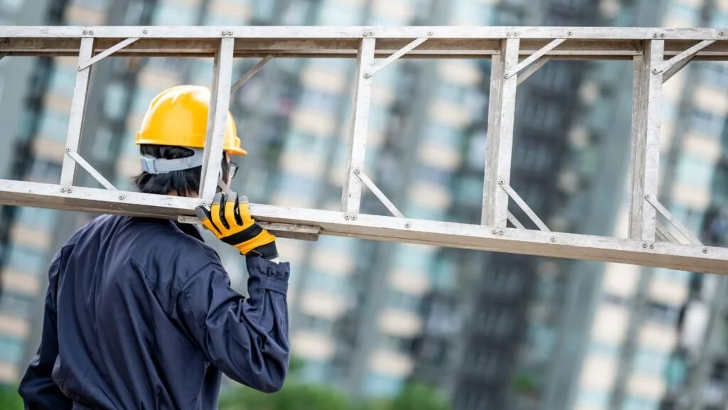 A construction worker wearing a yellow hard hat, blue jacket, and gloves carries a metal ladder at a construction site with apartment buildings in the background.