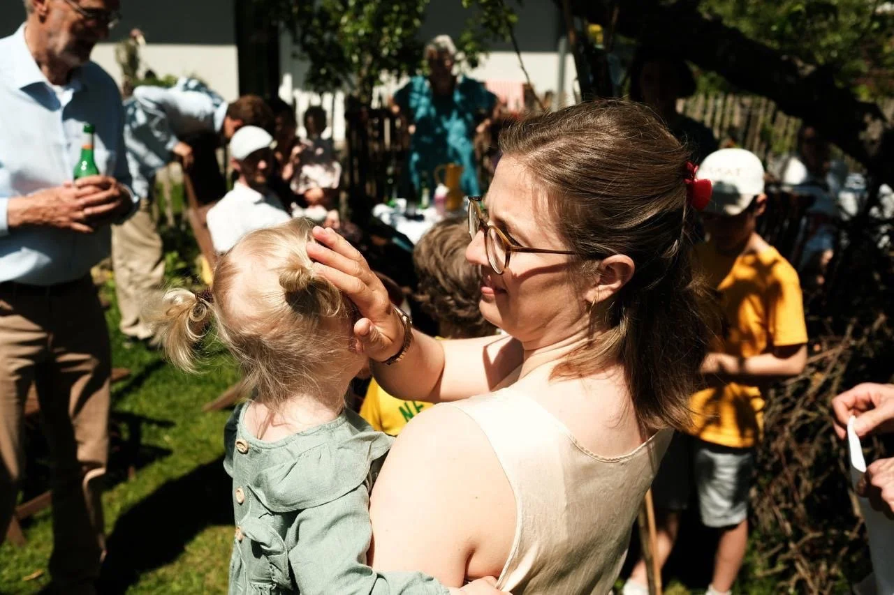 A woman with glasses holding a young girl in her arms at an outdoor gathering. The woman is gently touching the girl's head. Several people are in the background, seemingly enjoying a sunny day.