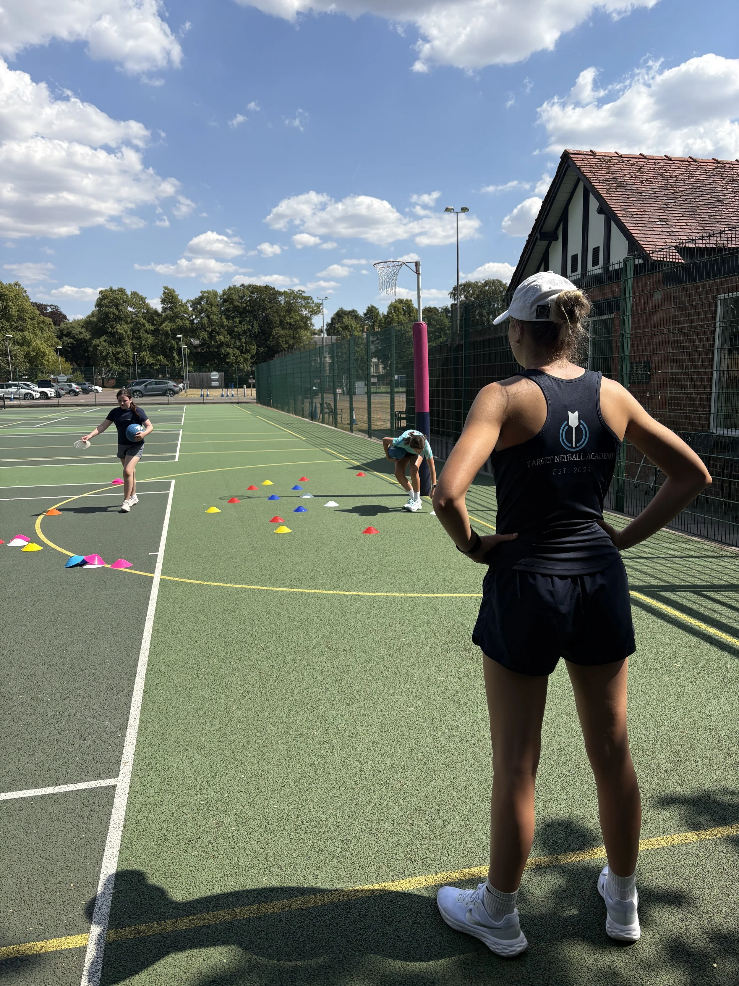 A woman with a white cap, black tank top with the Target Netball Academy logo, black shorts, and white sneakers watching children play netball on an outdoor court during the day.