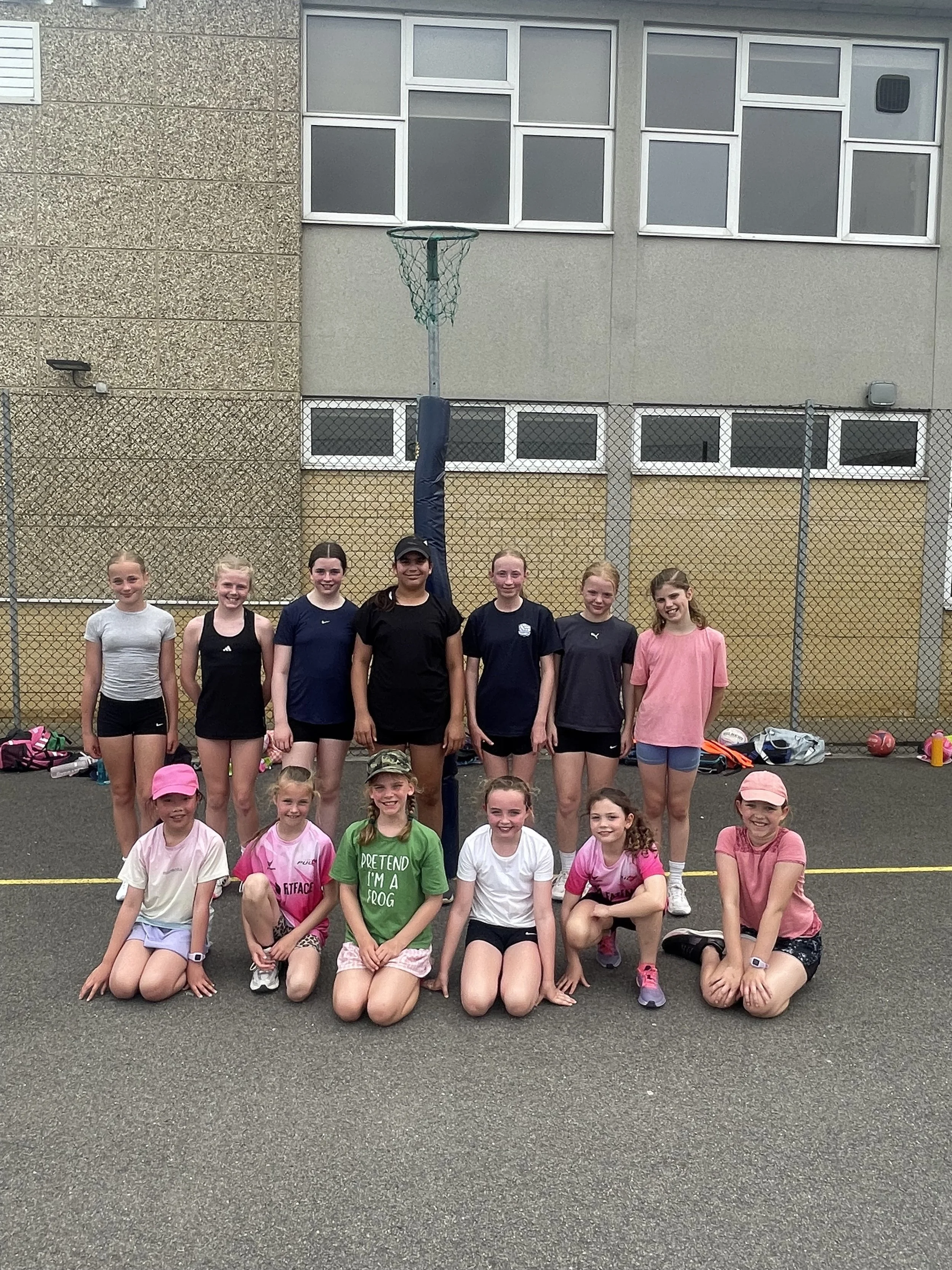 Group of young girls and women standing on a tennis court in athletic clothing, some kneeling in front, smiling at the camera. There is a building with windows and a chain-link fence in the background.
