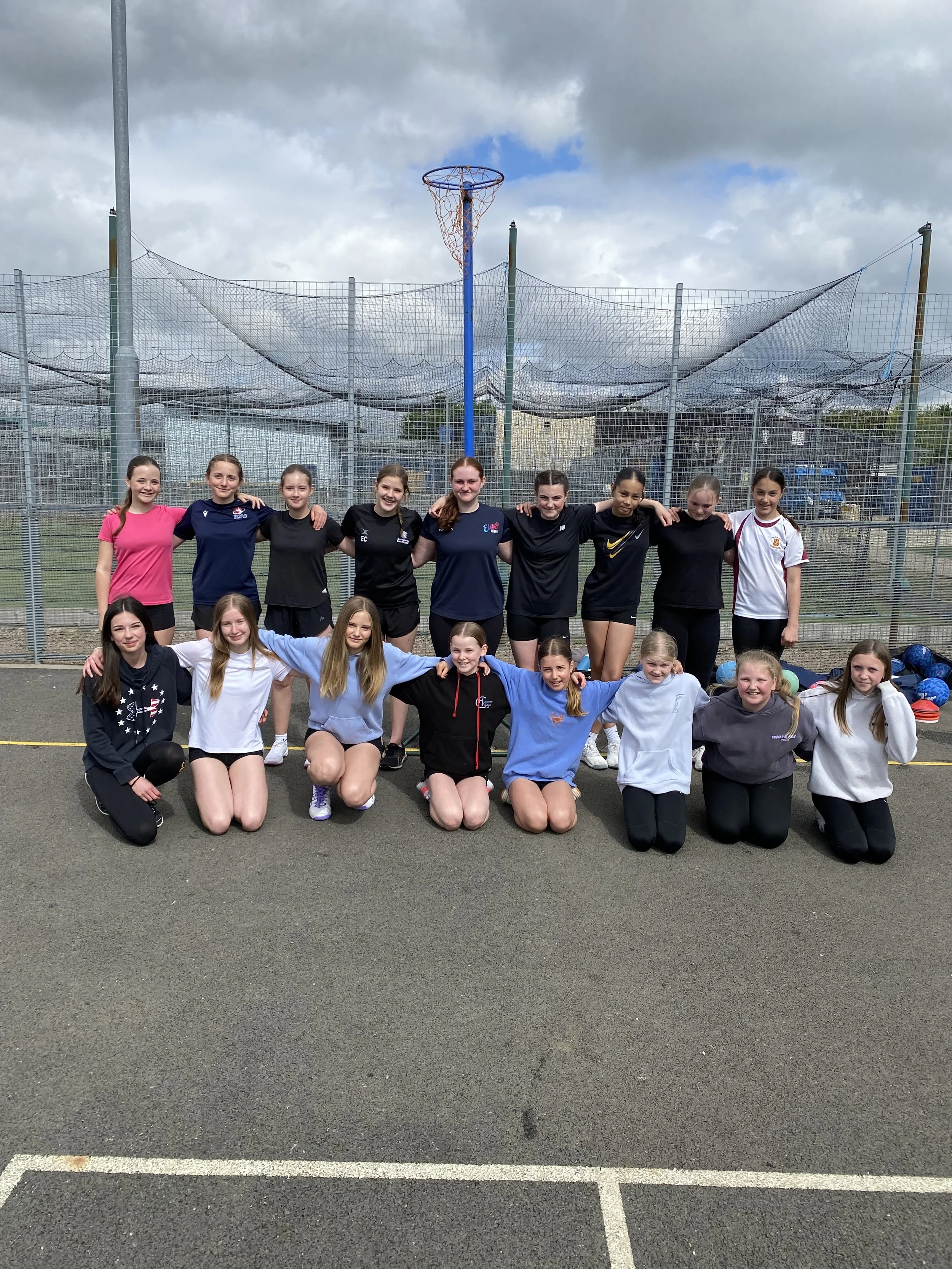 A group of young girls and women posing together on an outdoor basketball court. The group is standing with arms around each other, and some are kneeling in front. Behind them is a fence with a basketball hoop and cloudy sky.