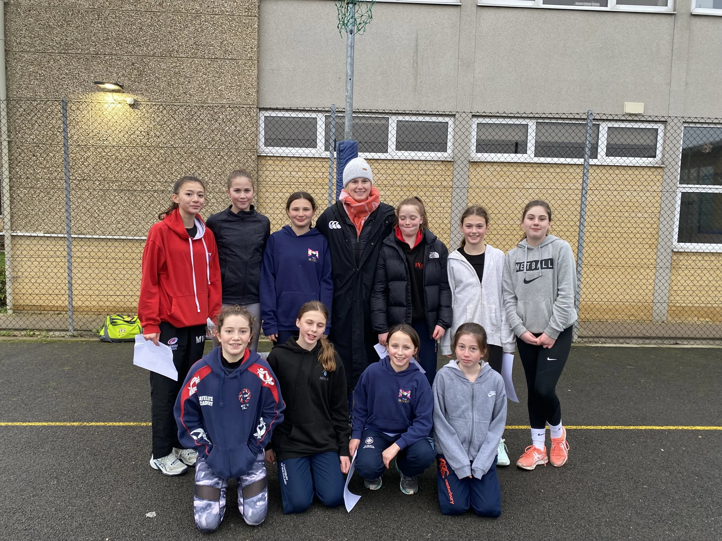 Group of young girls and their coach standing on a sports court with a chain-link fence and school building behind them, dressed in athletic clothing.