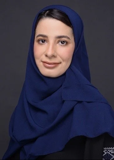 A woman wearing a blue hijab and black top, smiling softly at the camera against a dark gray background.