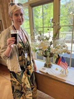 A woman holding a glass of wine standing next to a table with a floral arrangement and small American flag, by a window with green trees outside.