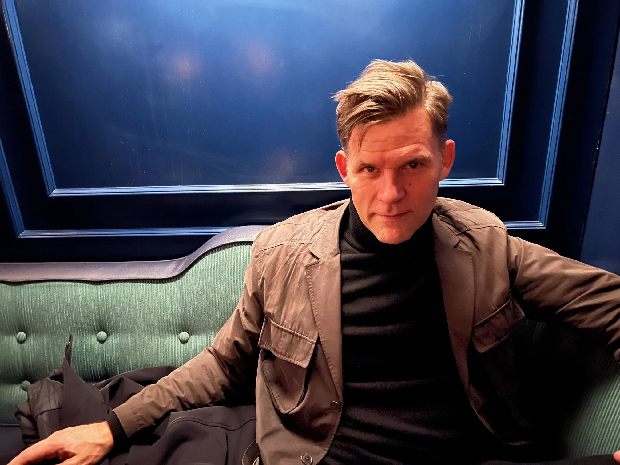 A man with blonde hair styled to the side, wearing a tan jacket over a black turtleneck, seated on a green upholstered bench in front of a blue wall with decorative trim.