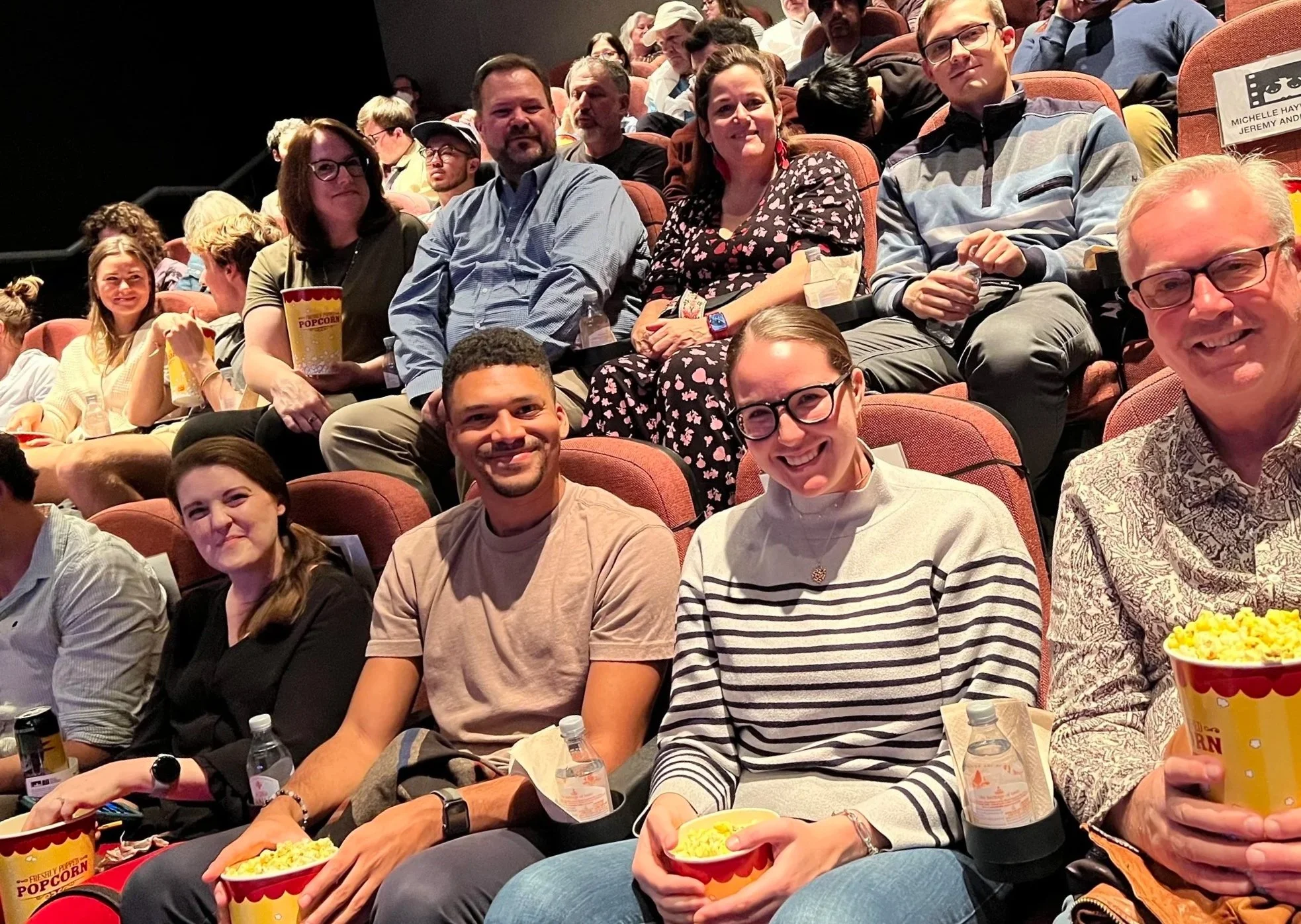 Audience members sitting in theater seats, watching a movie or event. People are smiling or focused on the screen, with some holding popcorn and drinks.