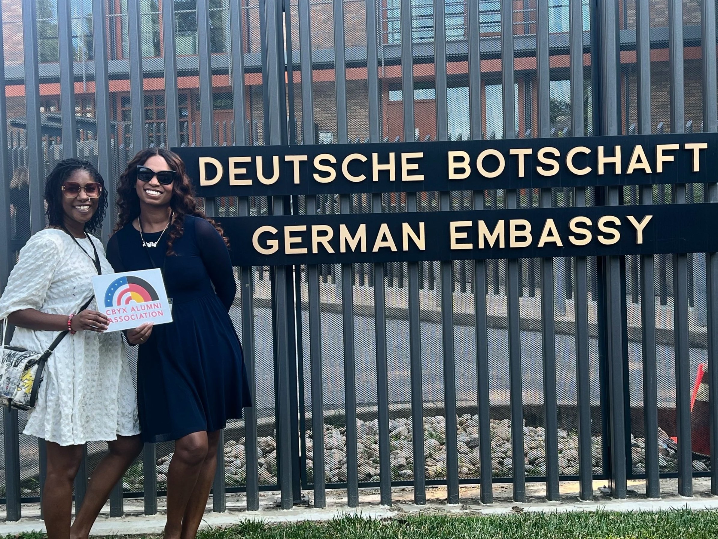 Two women standing outside near a gate with a sign for the German Embassy. One woman is holding a sign that says 'CBYX Alumnae Association' and they are both smiling.