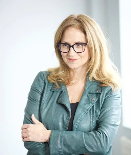 A woman with blonde hair and glasses wearing a teal leather jacket standing indoors with her arms crossed.