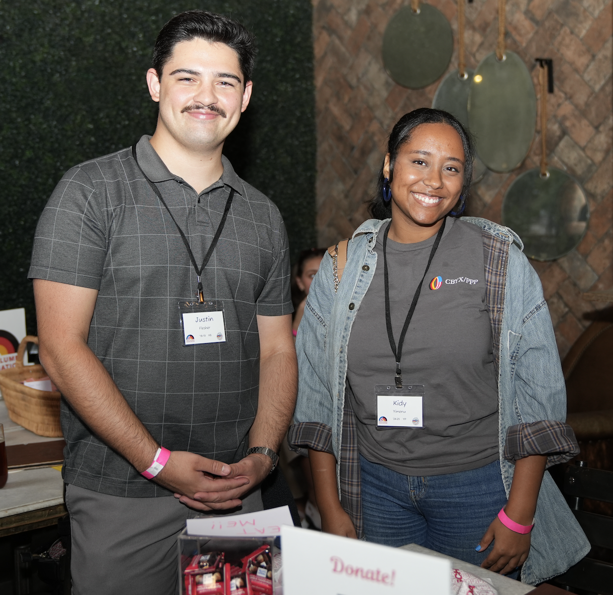 Two smiling young adults, a man and a woman, standing side by side at an indoor event with a brick wall and decorative round mirrors in the background. The man wears a gray checkered shirt, and the woman wears a gray t-shirt with a denim jacket, both