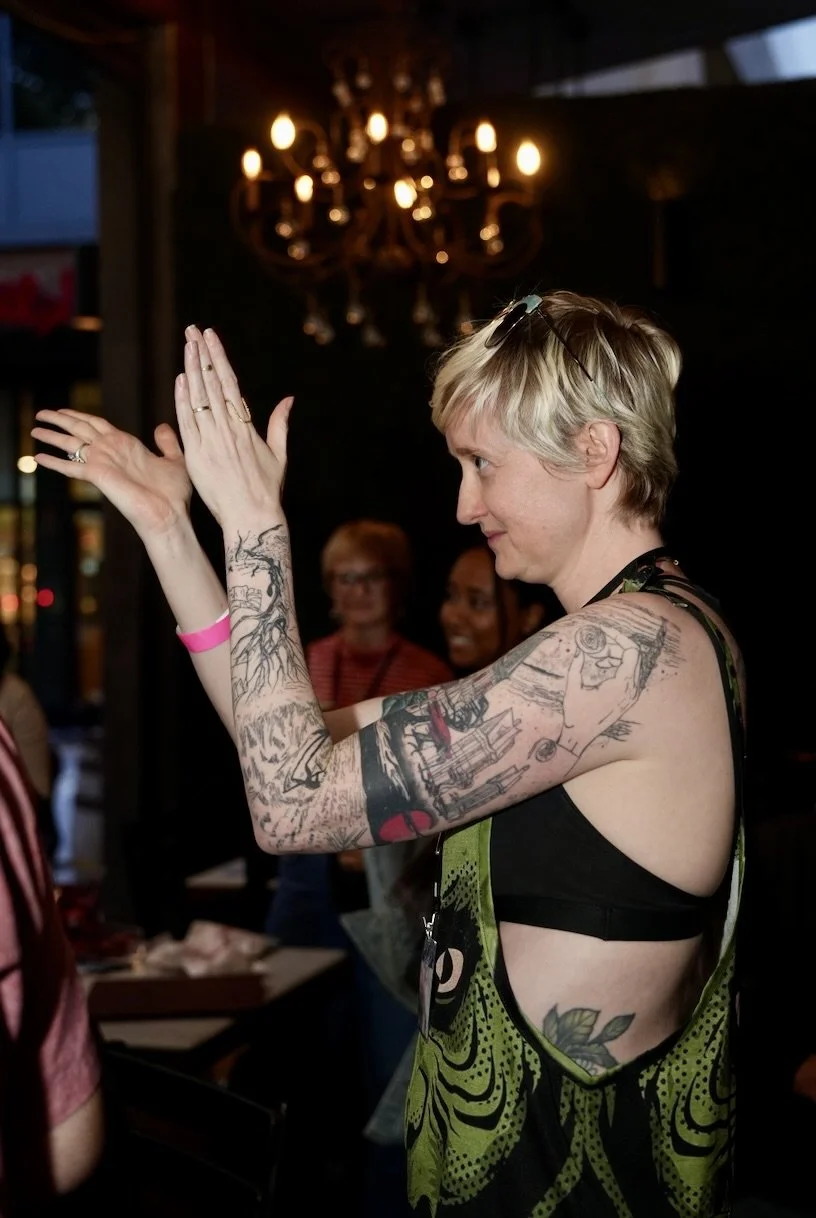 A woman with short blonde hair and tattoos on her arms, wearing a black top, is clapping her hands in a dimly lit room with a chandelier hanging above her.