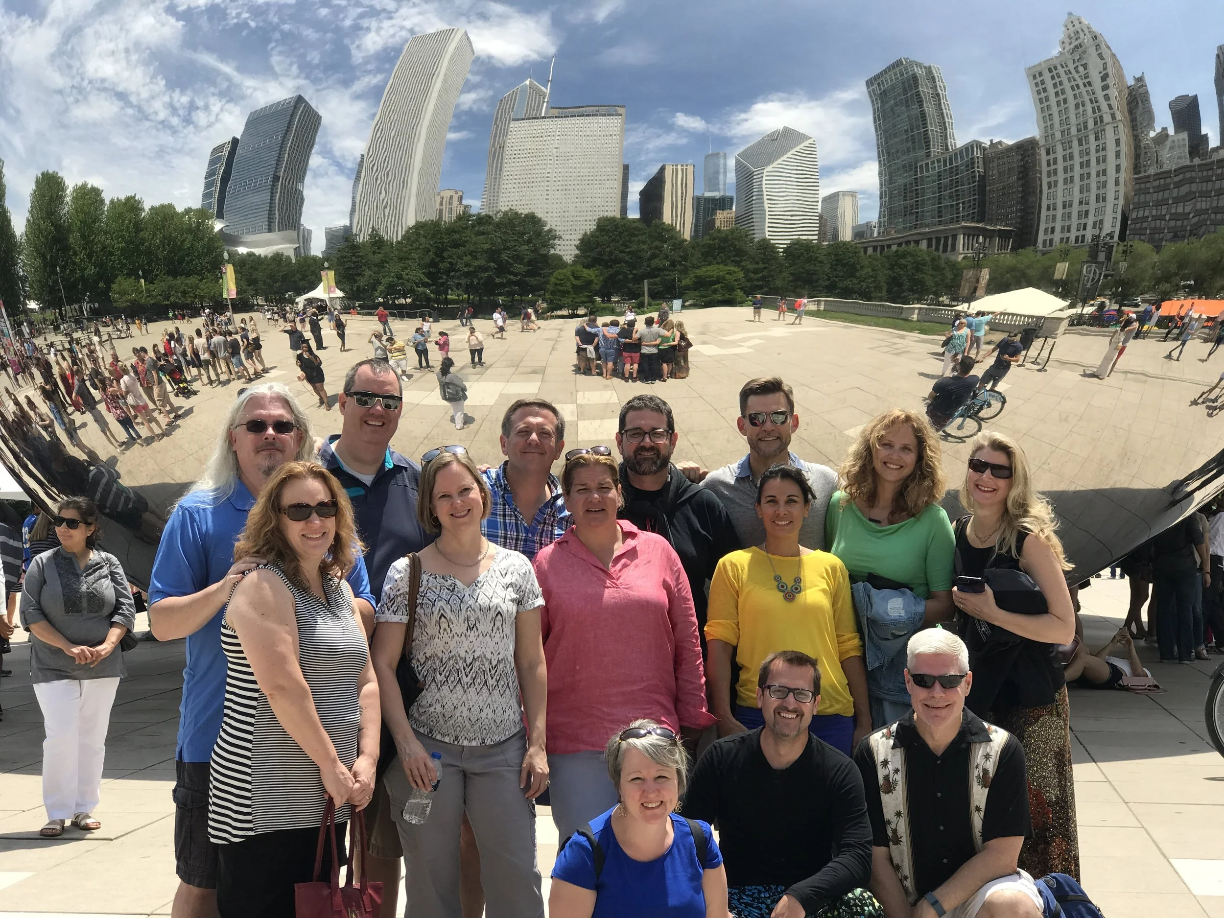 A group of diverse people posing for a selfie in front of a city skyline with tall modern buildings and a park with trees, under partly cloudy skies.