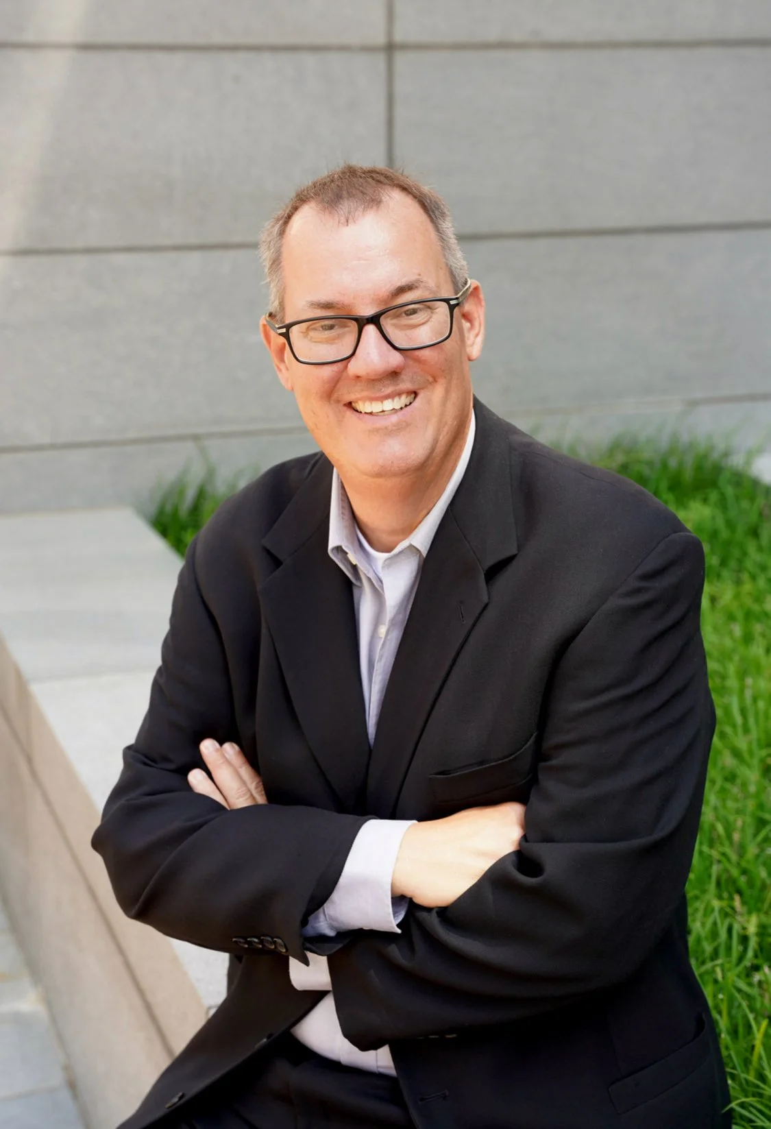 A man wearing glasses, a black suit, and a white shirt, smiling with arms crossed in an outdoor setting with concrete and green plants.