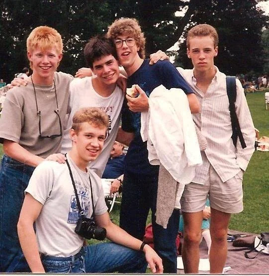 Five young boys posing together outdoors, smiling and looking at the camera, with trees and a grassy area in the background.