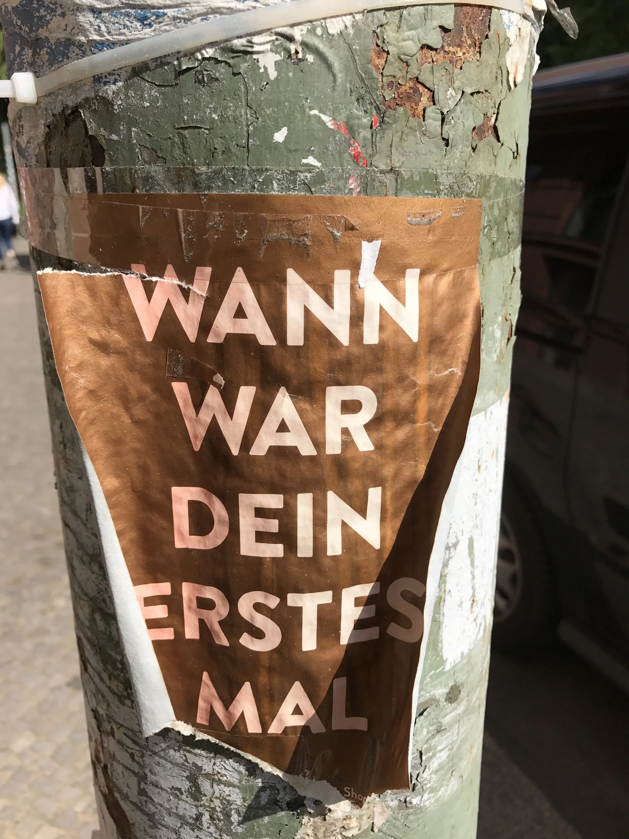 Close-up of a weathered, peeling poster on a metal pole with German text that reads "WANN WAR DEIN ERSTES."
