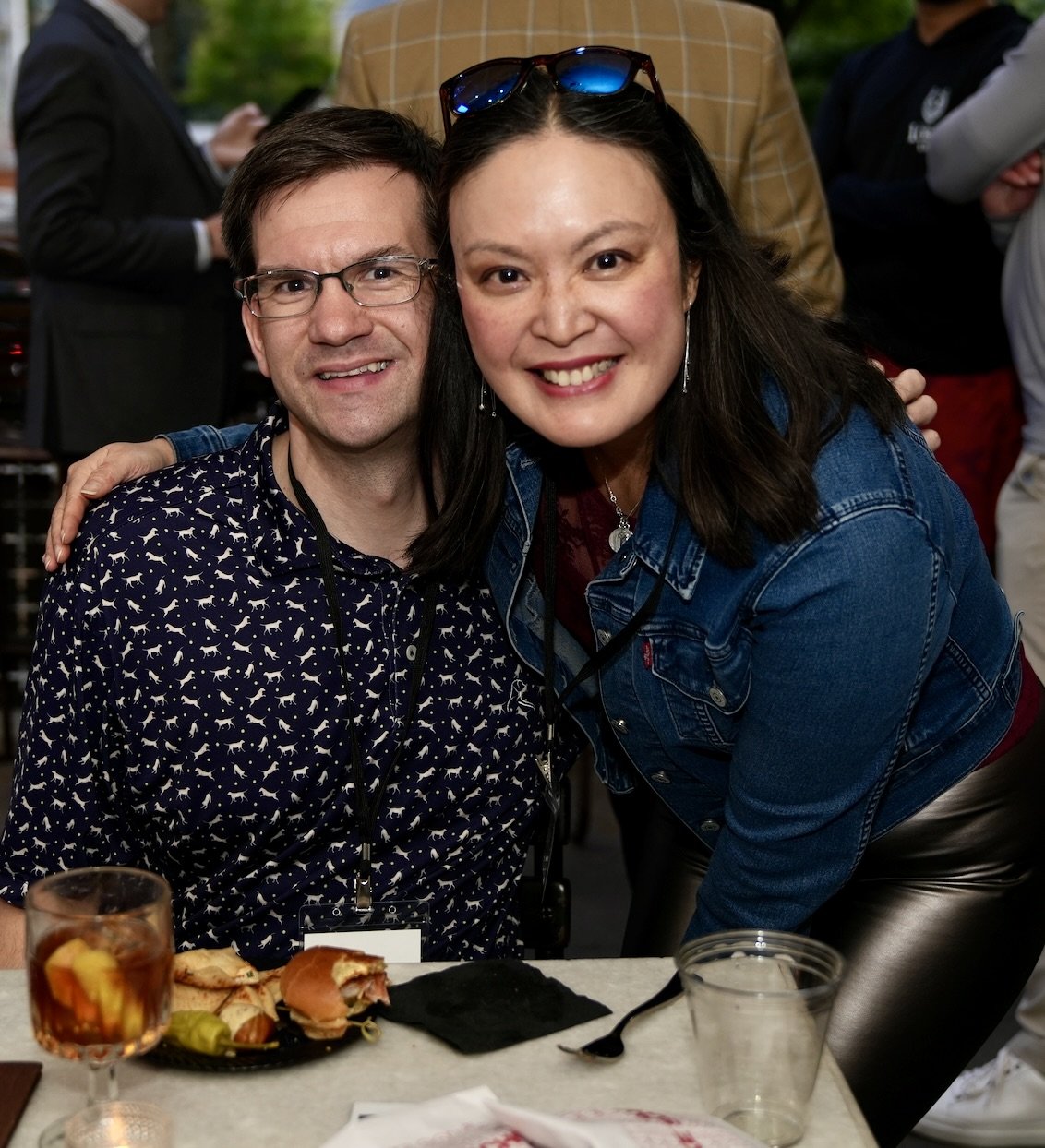 Two people smiling and posing together at a social gathering, seated at a table with food and drinks, with other guests in the background.