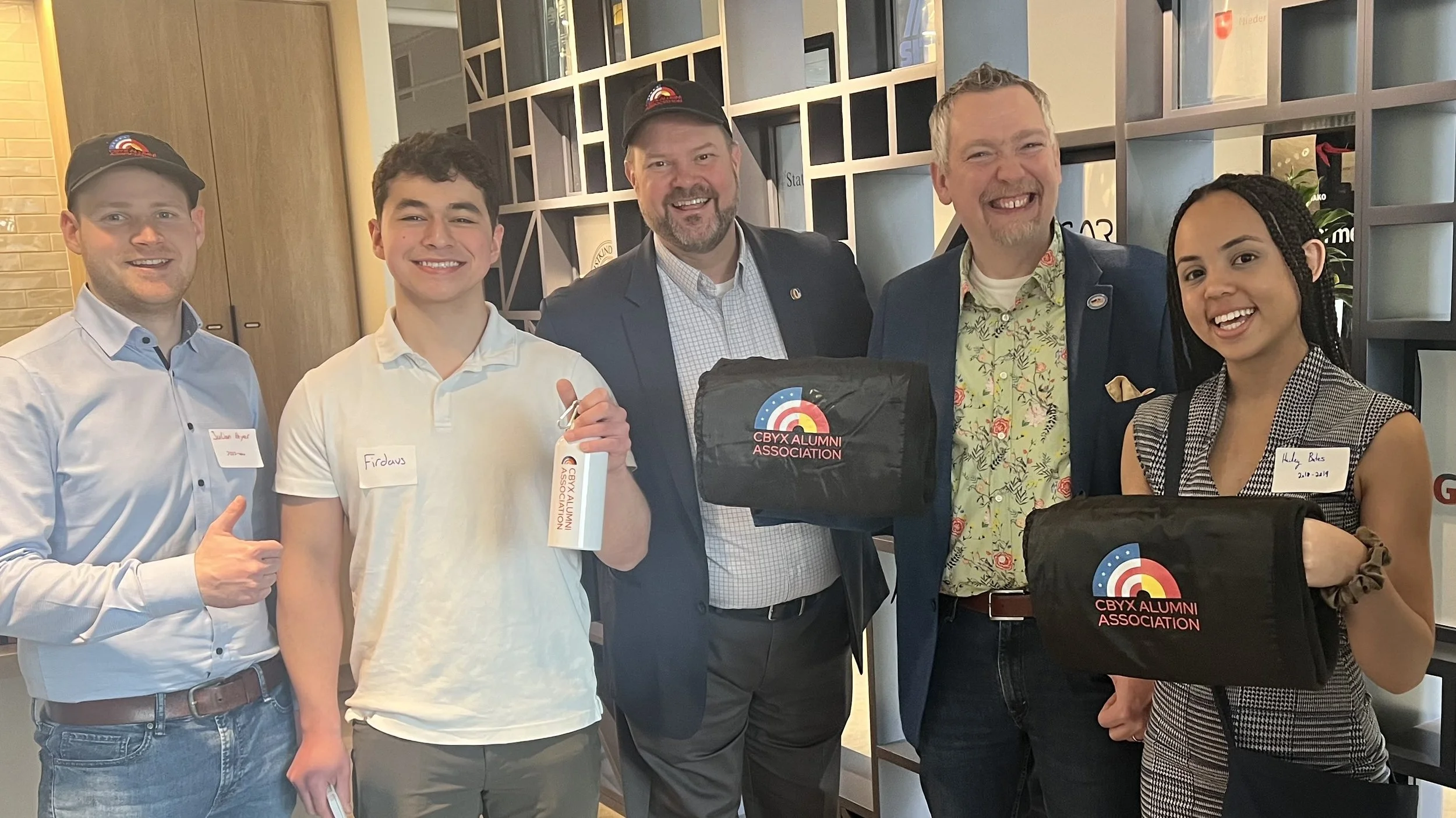 Group of five diverse people smiling, holding CBYX Alumni Association bags at an indoor event.