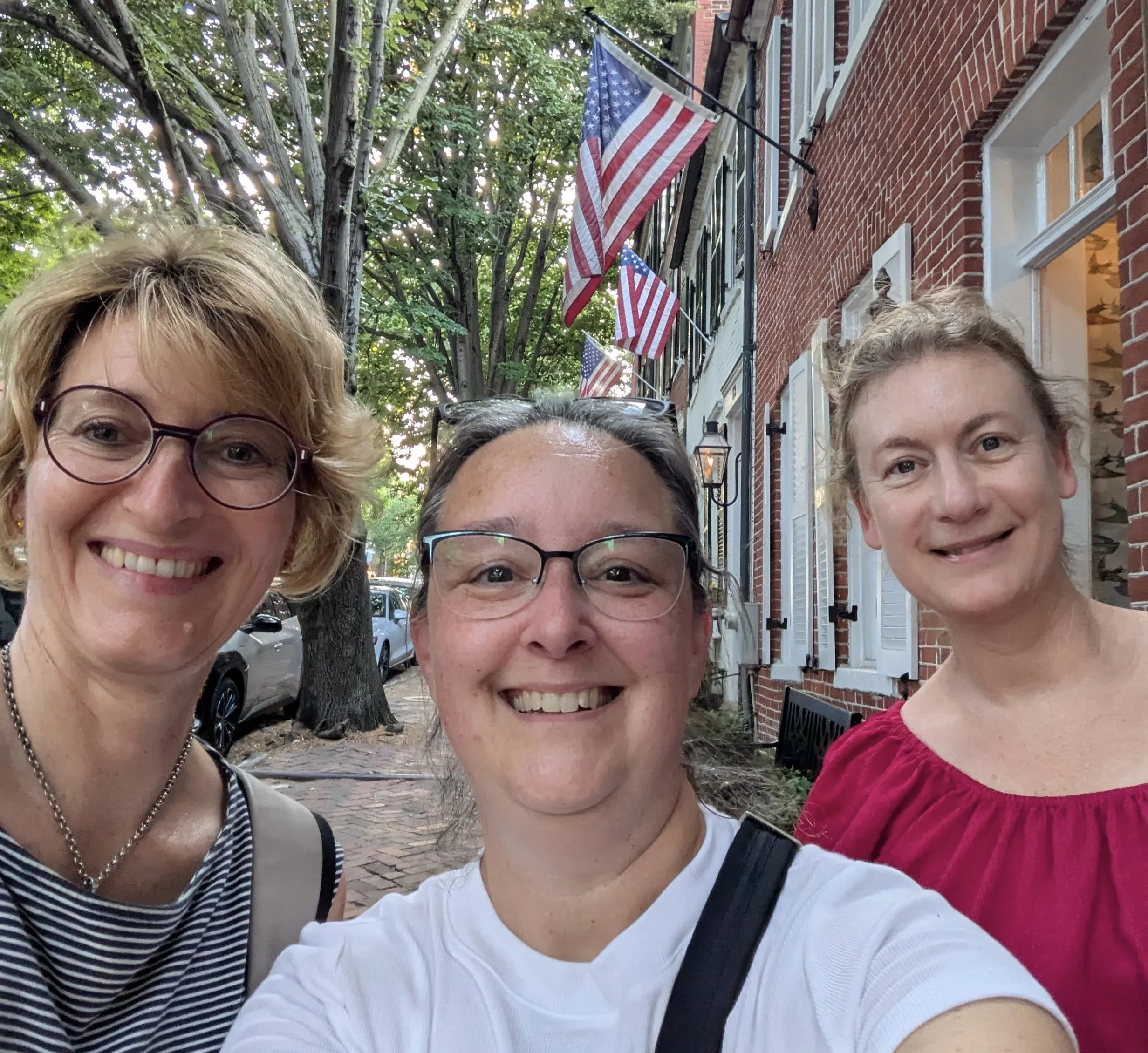 Three women taking a selfie outdoors on a city street with American flags hanging above and trees in the background.