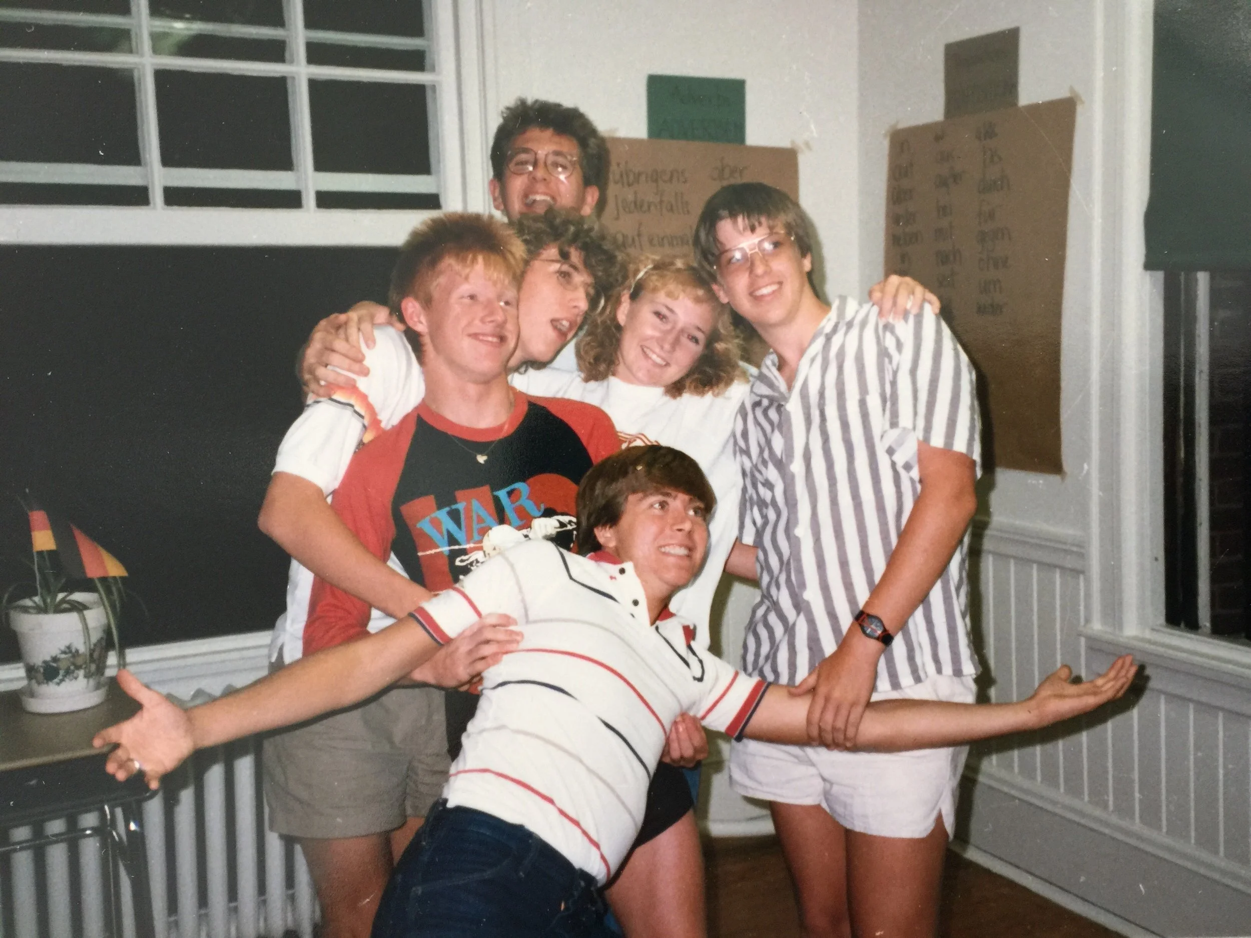Group of five young people posing together indoors. They are smiling, and some are hugging or holding each other. The background includes windows, a green chalkboard, and cardboard signs on the wall.