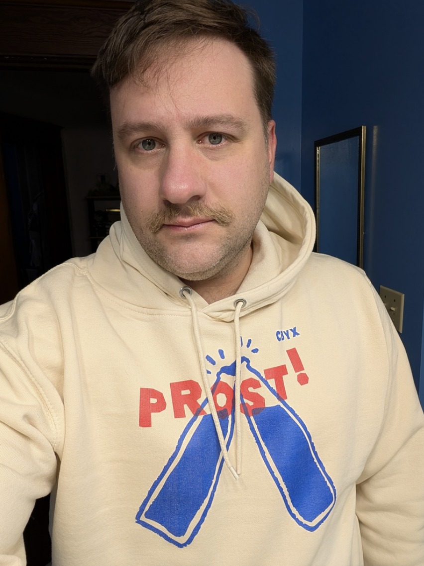 A man taking a selfie indoors, wearing a beige hoodie with red and blue graphics and text that says 'PROTEST!' with two blue bottles. The background shows a blue wall and a framed picture.