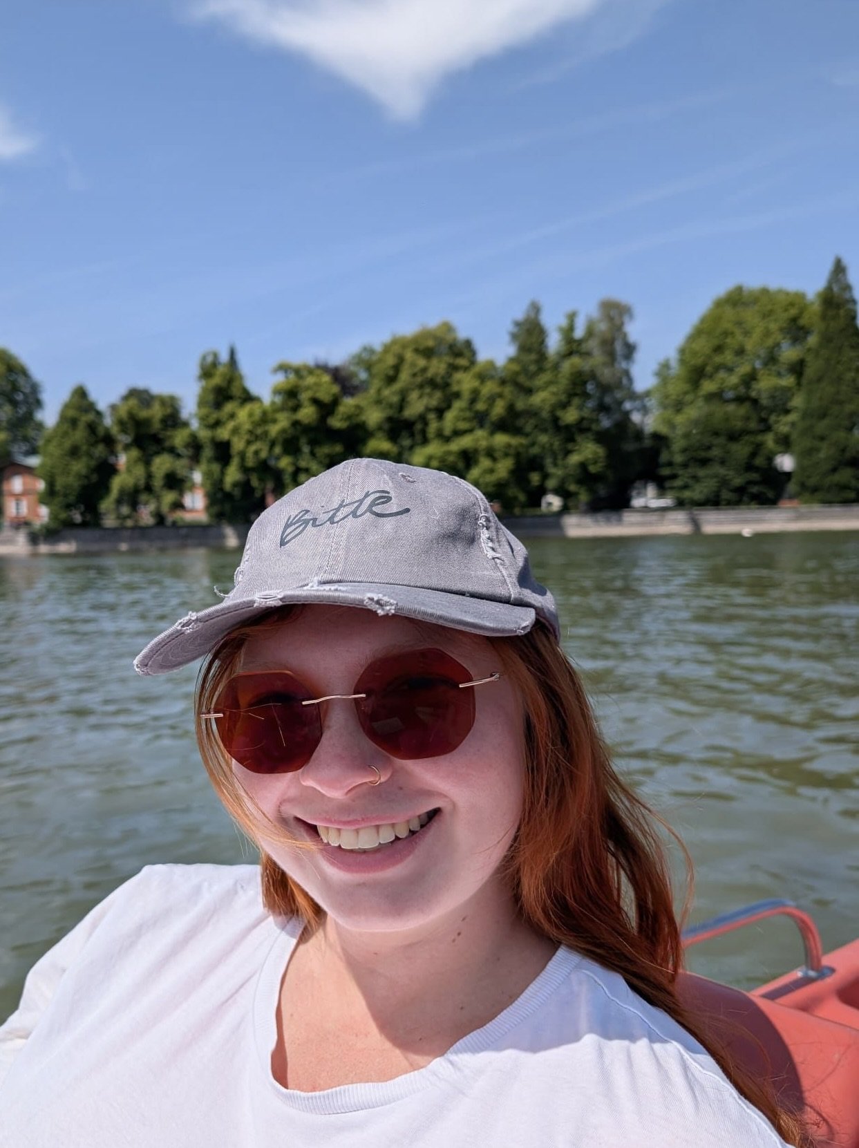 A woman with red hair wearing round sunglasses and a gray cap smiling in front of a river, with green trees and buildings in the background on a sunny day.
