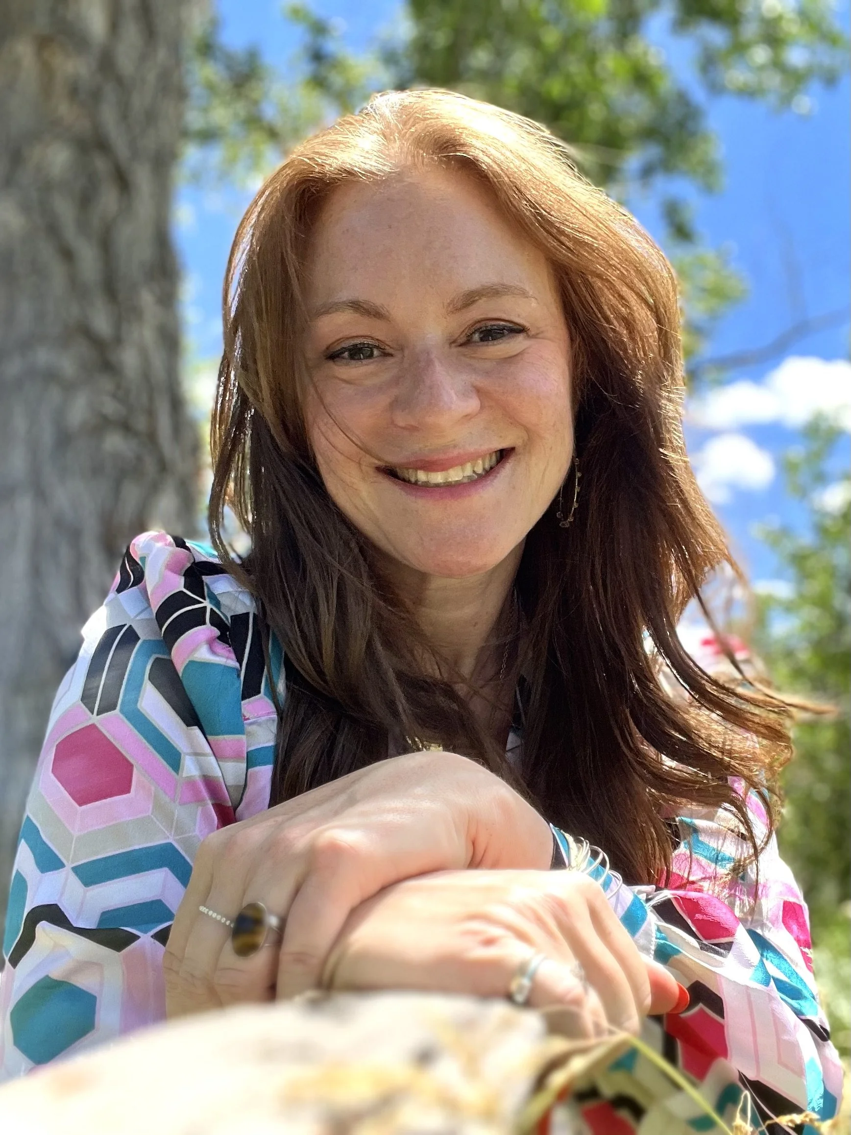 Close-up of a smiling woman with reddish-brown hair, outdoors on a sunny day with blue sky and green leaves in the background.