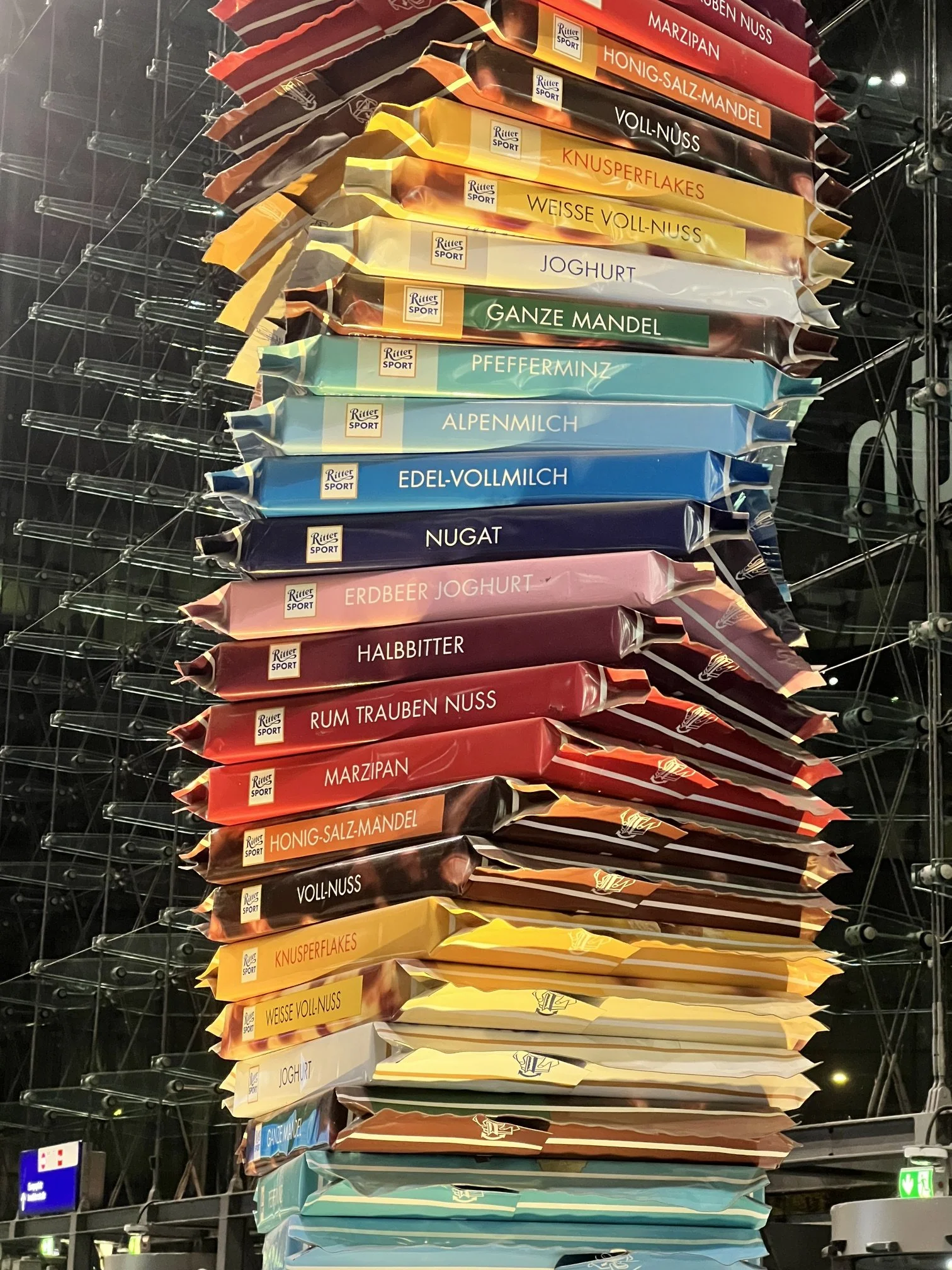 Stacked packages of Ritter Sport chocolate flavors, including Marzipan, Honig-Salz-Mandel, Voll-Nuss, Knusperflakes, Weisse Voll-Nuss, Joghurt, Ganze Mandel, Pfefferminz, Alpenmilch, Edel-Vollmilch, Nugat, Erdbeer Joghurt, Halbbitter, Rum Trauben Nuss, and Marzipan, are arranged vertically on a display stand.