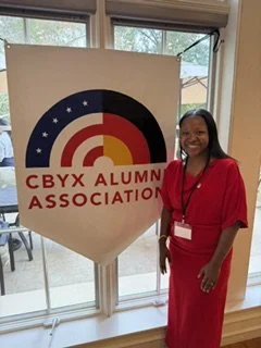 A woman in a red dress standing next to a CBYX Alumni Association banner inside a room with large windows. Halle Foundation. 