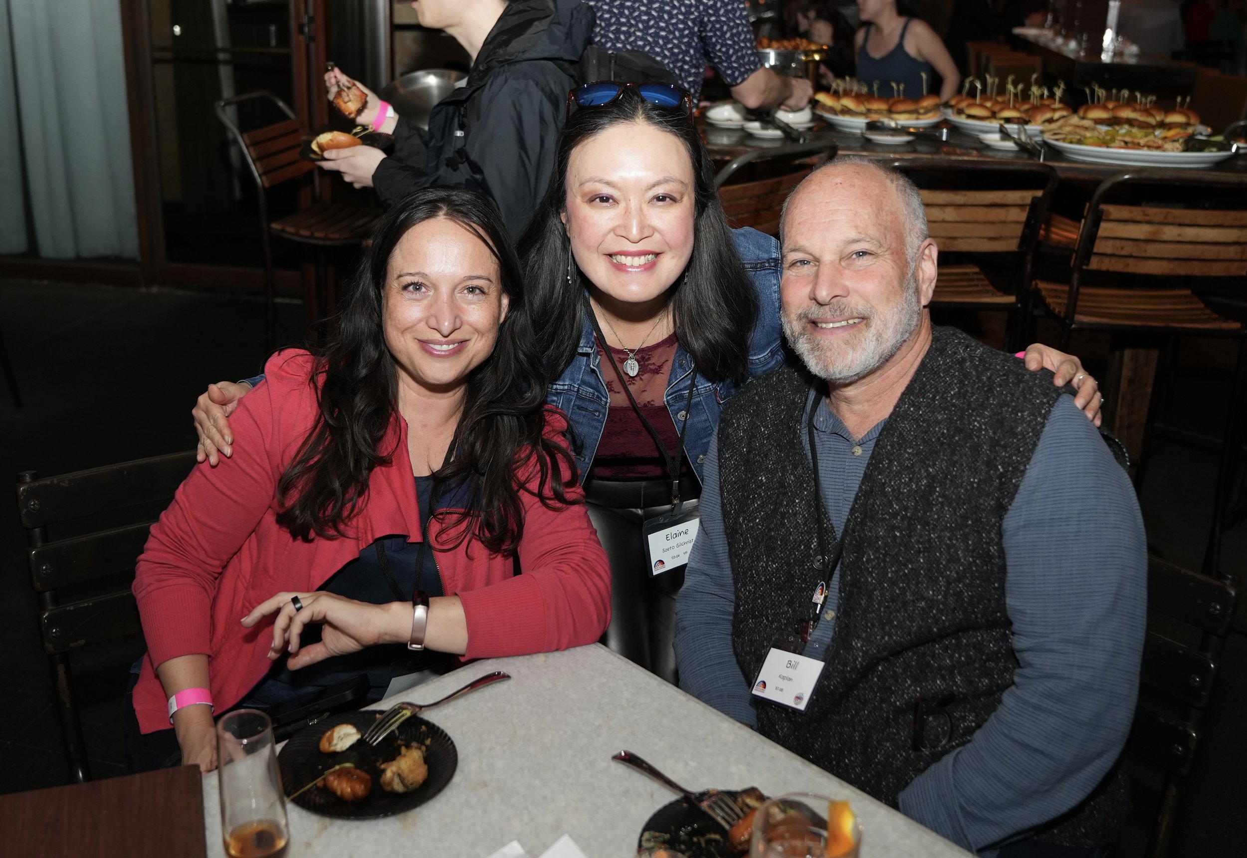 Three people smiling at a table with food in a restaurant or event setting. A woman in a red top sits on the left, a woman with dark hair and sunglasses on her head in the middle, and a man with a gray beard on the right. In the background, there are