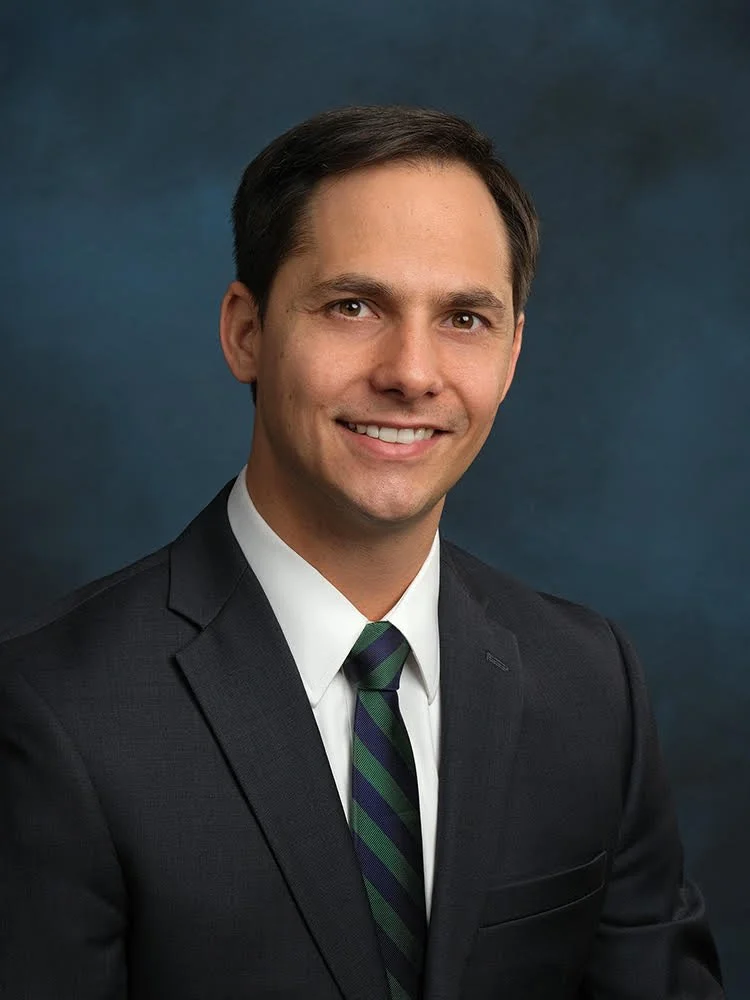 A professional headshot of a man with short dark hair, wearing a black suit, white shirt, and a navy blue and green striped tie, smiling against a dark blue background.