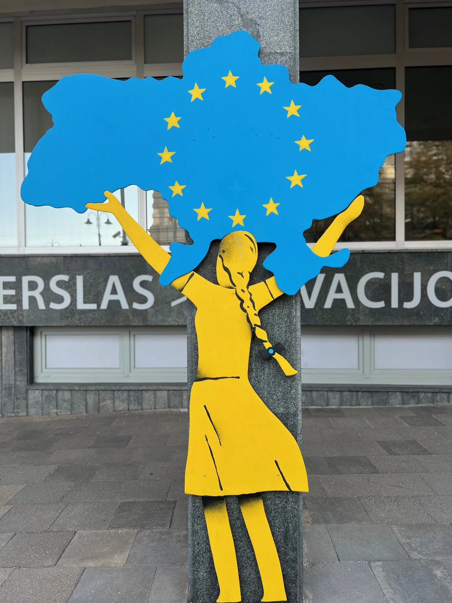 A colorful cut-out figure of a woman with blue hair shaped like the European Union flag, yellow arms, and yellow legs, standing outdoors against a gray wall with large windows.