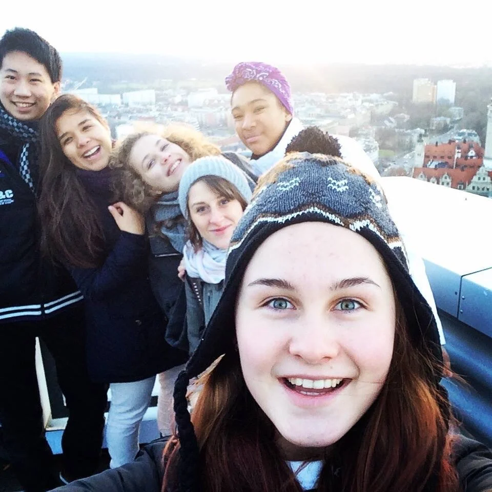 Group of six young friends taking a selfie on a rooftop with a cityscape background at sunset.