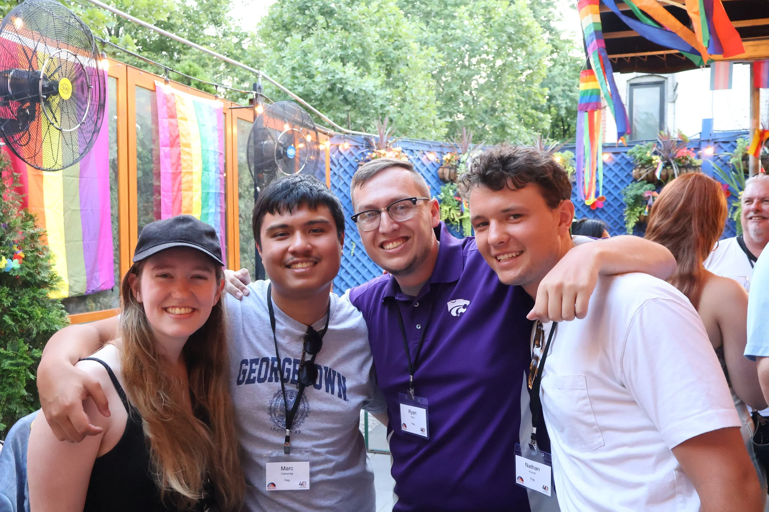 Group of young people smiling and posing together at an outdoor party decorated with rainbow flags and pride flags, with trees and string lights in the background. National Reunion, Washington, DC, Prost, CBYX Alumni,