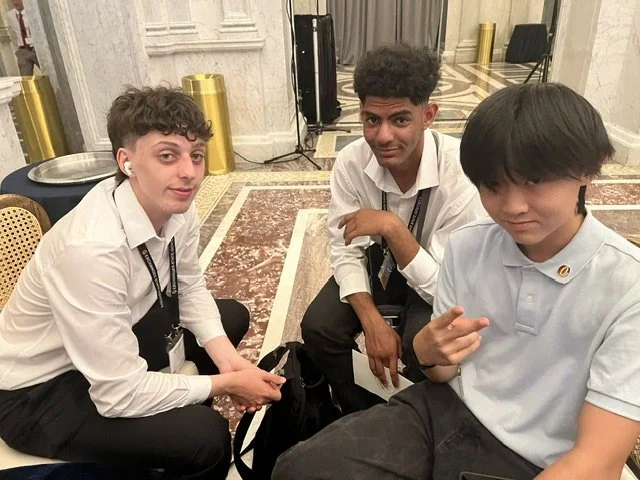 Three young men sitting and posing for a photo indoors, with a ornate carpet, gold-colored decorative elements, and a reflective surface in the background.