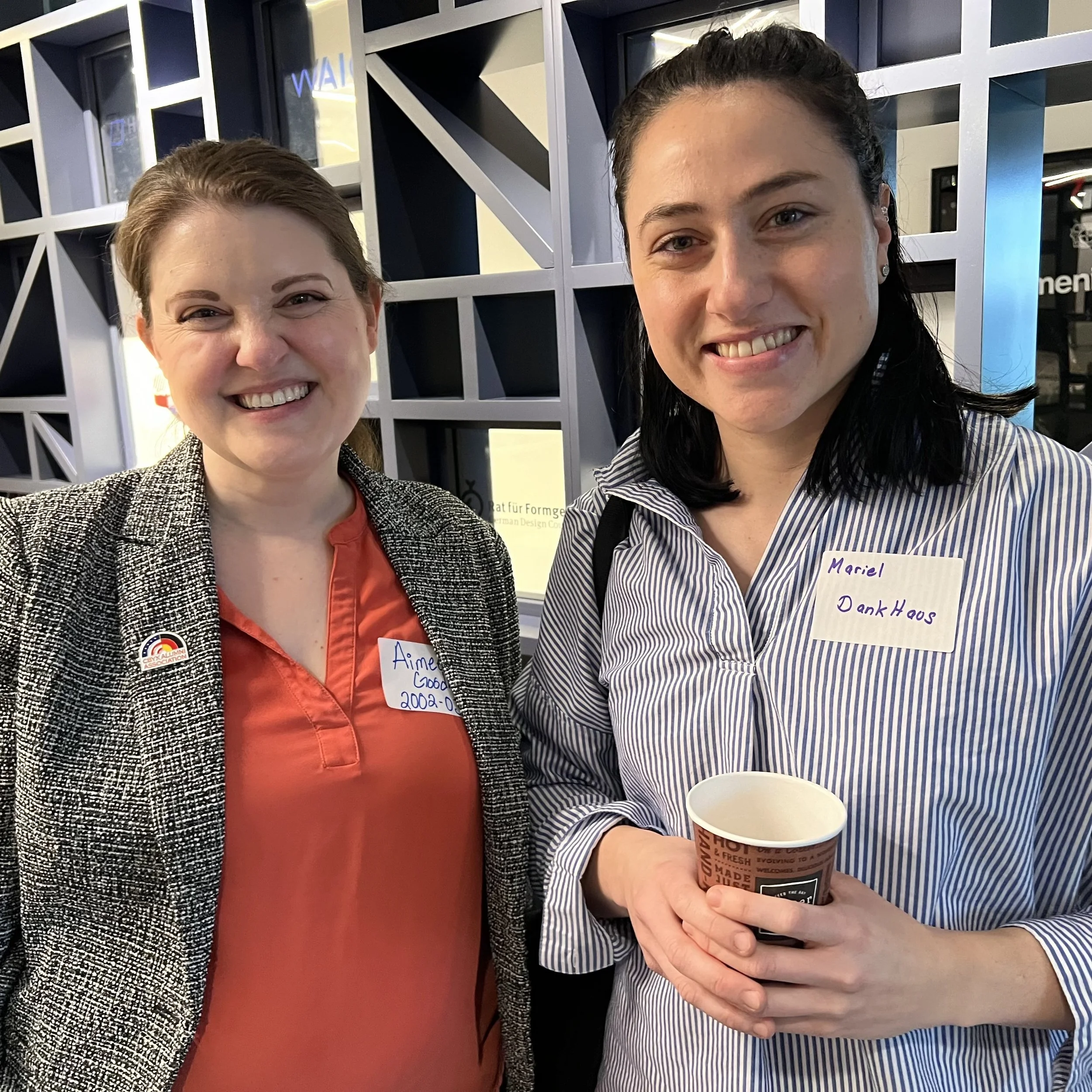 Two women smiling for a selfie in front of a geometric patterned wall inside a building, one holding a coffee cup.