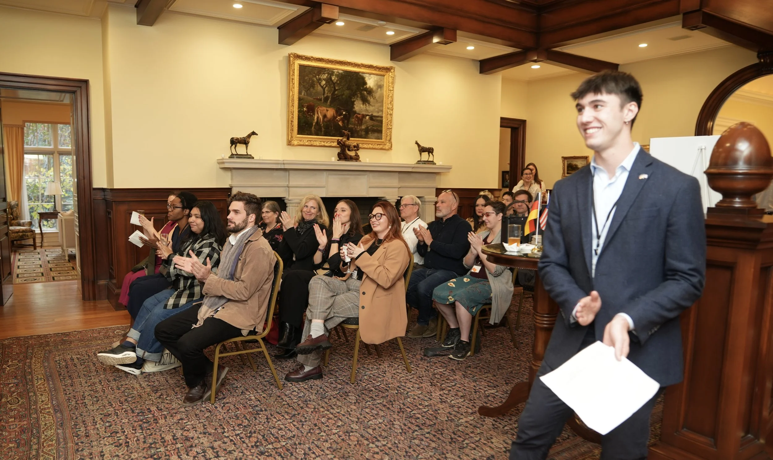 People seated and clapping in a formal room with ornate decorations, paintings, and wooden beams, while a young man in a suit stands in the foreground smiling.