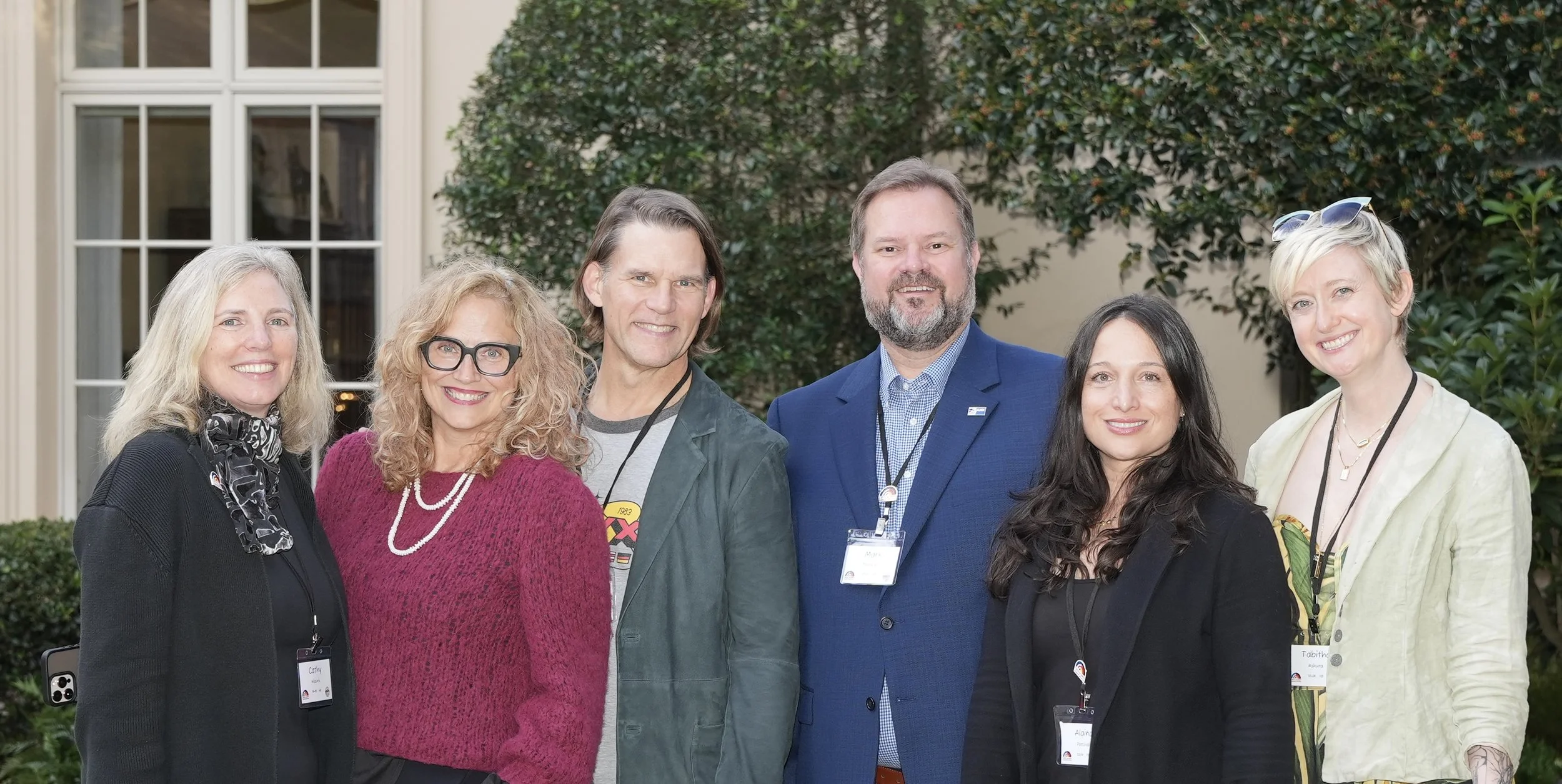 Group of six diverse people standing outdoors in front of a building with green bushes, smiling at the camera.