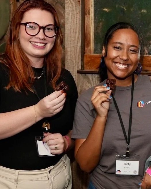 Two women smiling and holding pieces of chocolate, standing in a room with wooden walls and a window.