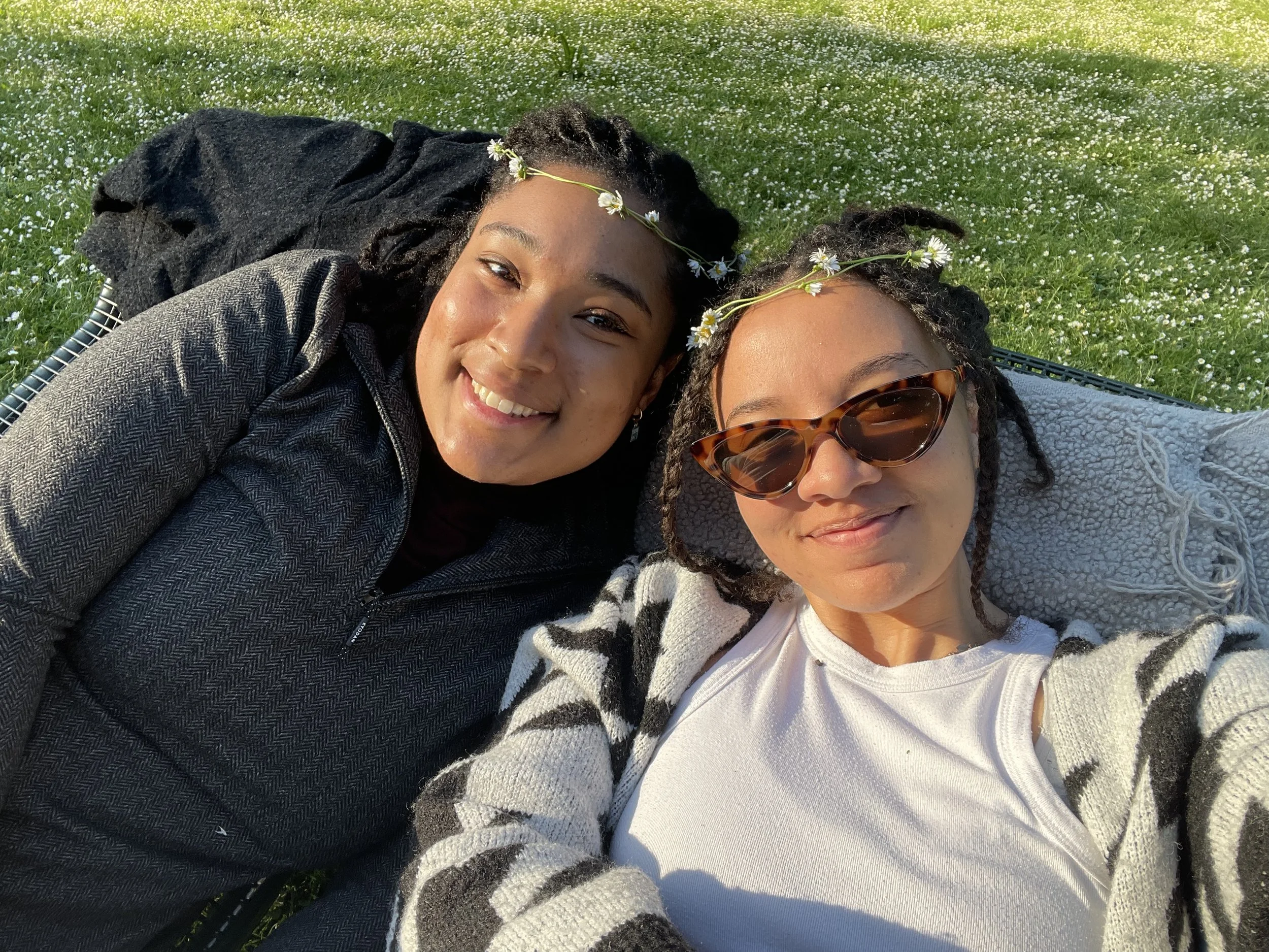 Two women lying on grass in sunlight, smiling, wearing flower crowns, one with sunglasses, relaxed outdoor scene. CBYX Alumni, Young Professionals. 