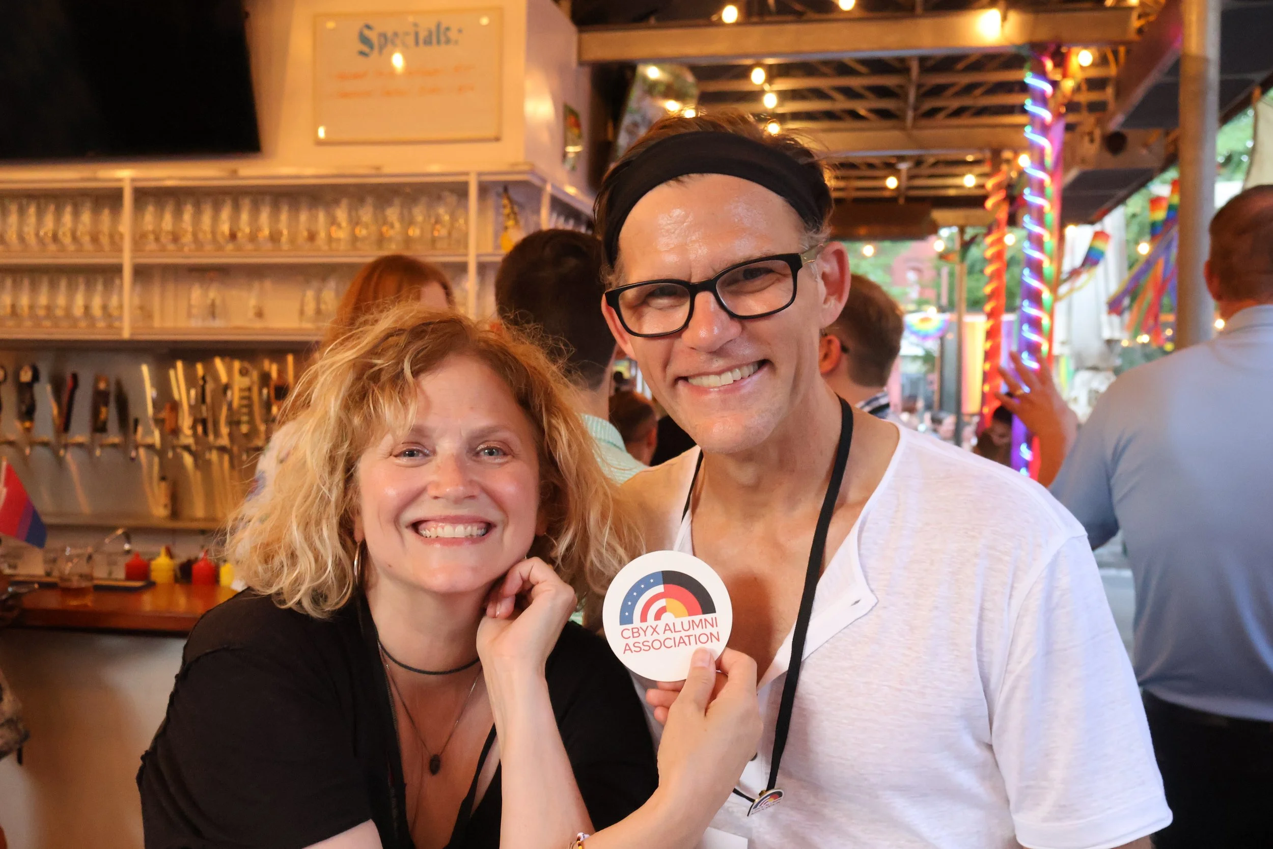 Two smiling women and a smiling man at a festive event, with the men holding a CBYX Alumni Association badge, in an outdoor setting with string lights and colorful decorations.