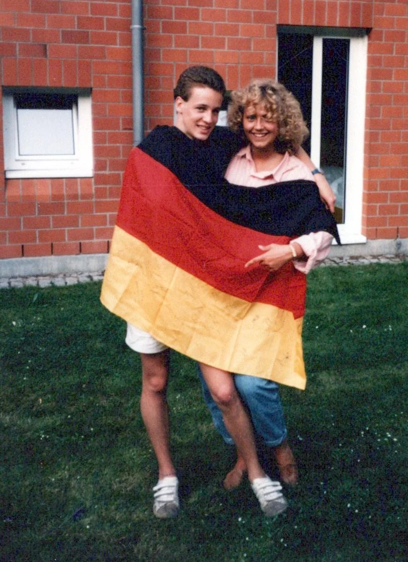 Two people standing on grass in front of a brick building, wrapped together in a German flag, smiling at the camera.